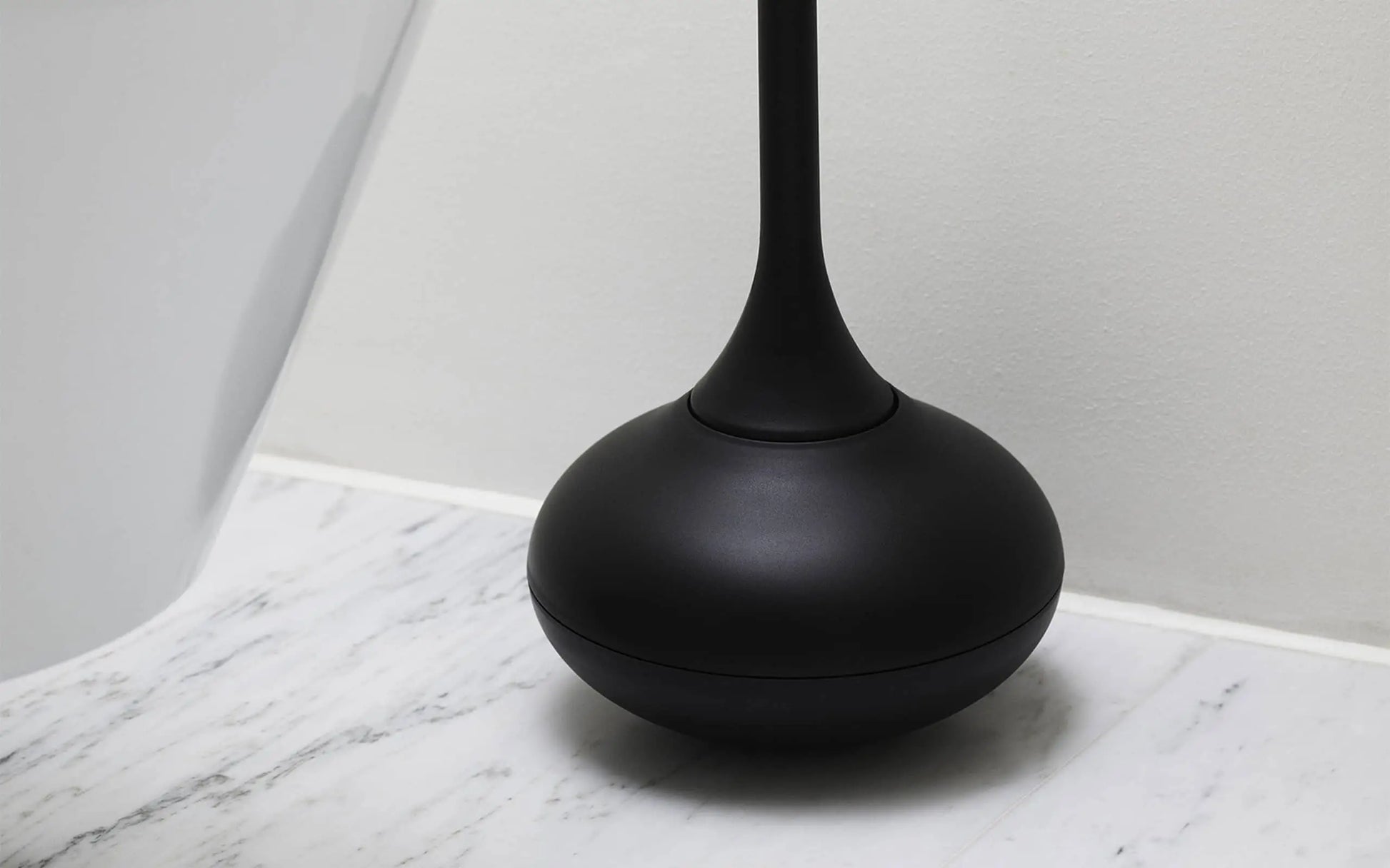 The Normann Copenhagen Ballo Toilet Brush, with its rounded base and modern design, sits on a white marble bathroom floor beside a toilet. A smooth, light wall behind adds style to your bathroom cleaning routine.