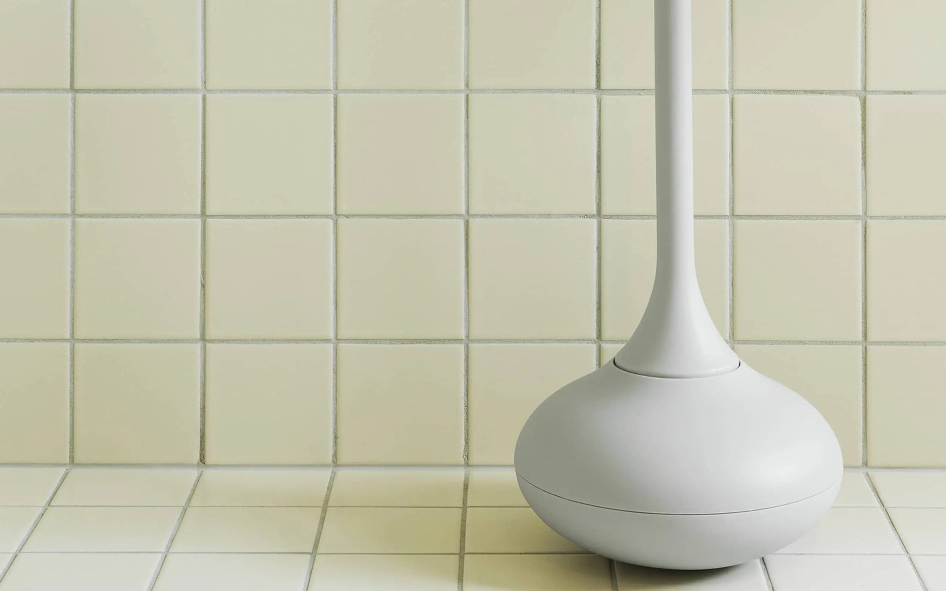 The Normann Copenhagen Ballo Toilet Brush, in white, features a rounded base and stands on a tiled bathroom floor beside a wall of square pale yellow tiles.