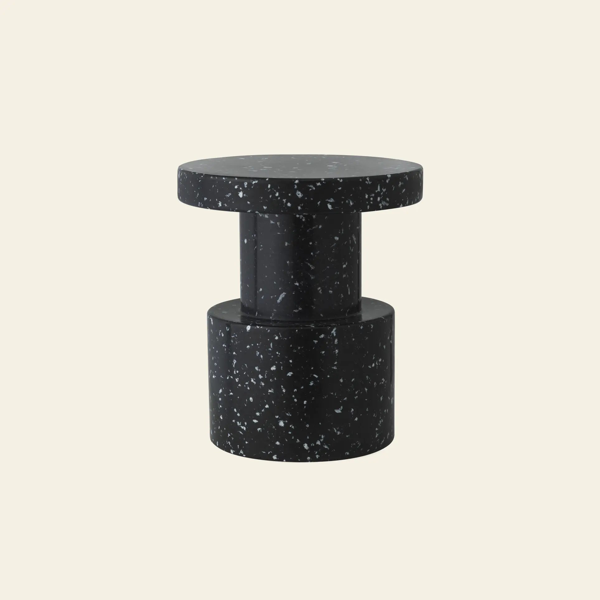 The Normann Copenhagen Bit Stool, a compact modern black seat with white speckles, features a round top and cylindrical base, highlighting its multifunctional design against a plain off-white background.