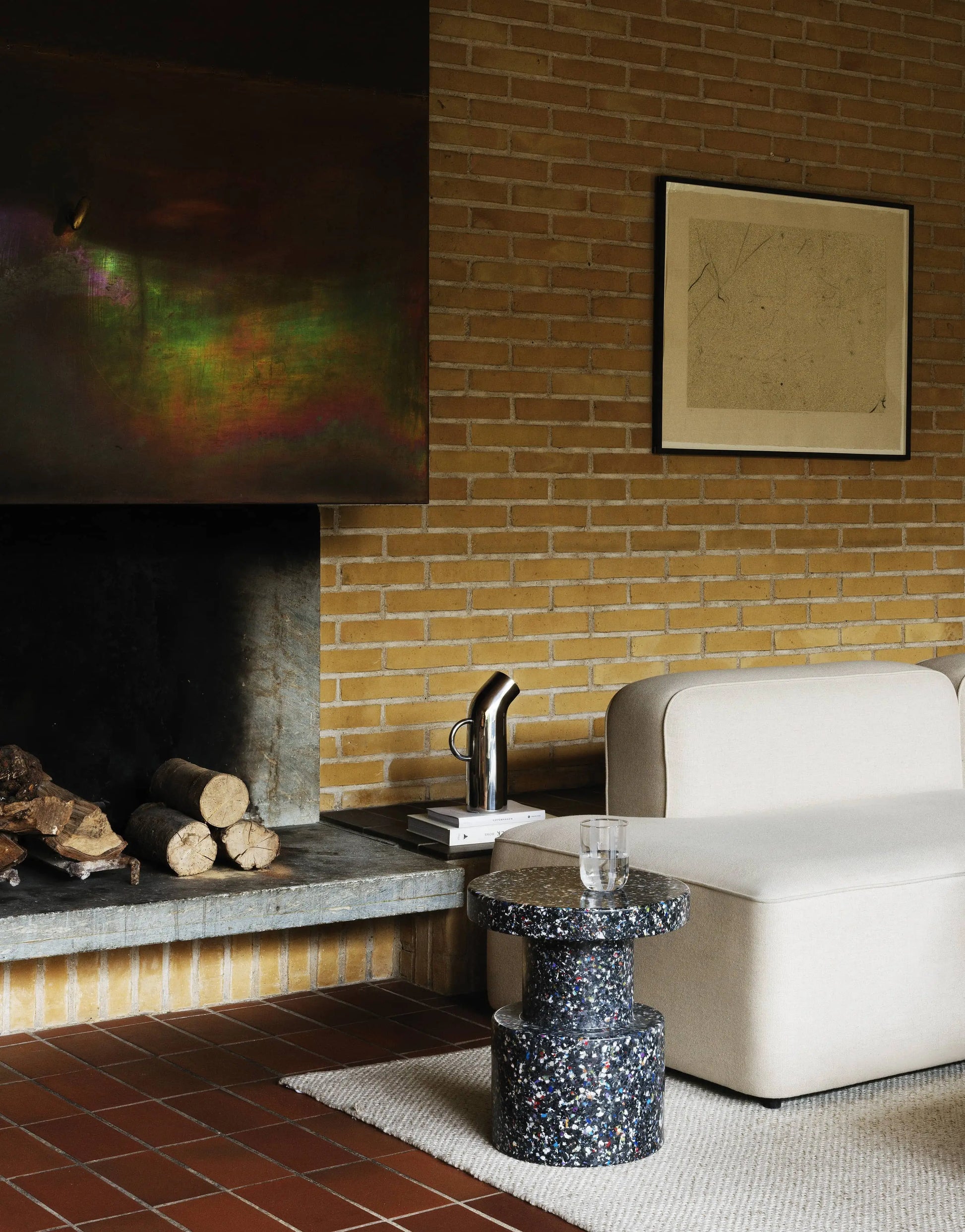A modern living room with a beige sofa, a Normann Copenhagen Bit Stool as a speckled side table holding a glass, a fireplace with stacked logs, and brick walls adorned with abstract minimalist art.