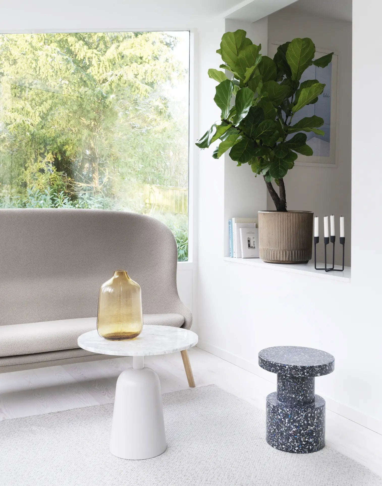 A modern living room with a beige sofa, a round white marble coffee table holding a large amber glass vase, a terrazzo accent table, extra seating from the Normann Copenhagen Bit Stool, a potted fiddle leaf fig, and a window with greenery outside.