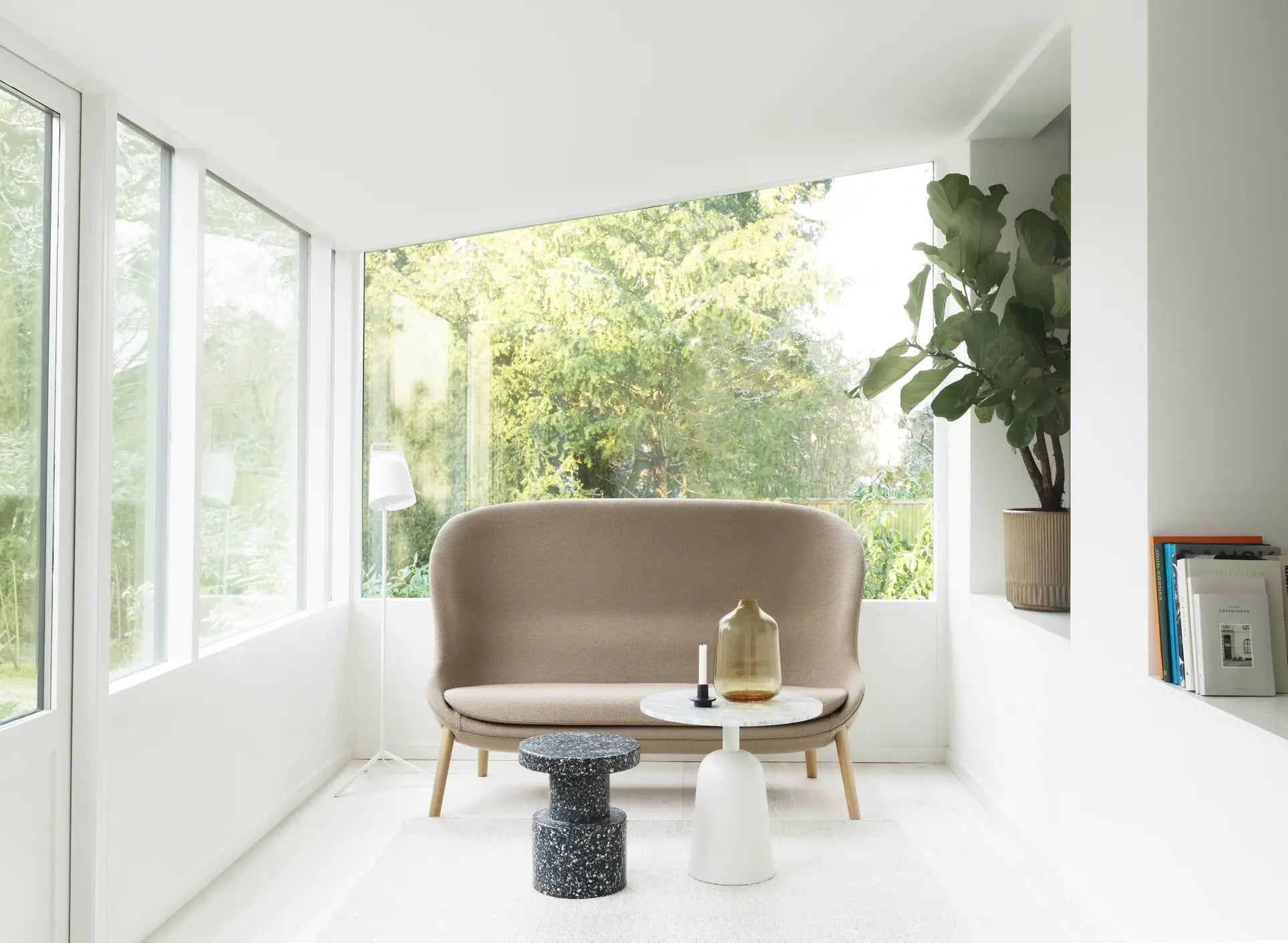 A bright, minimalist living room with large windows, a beige loveseat, versatile seating like the Normann Copenhagen Bit Stool, two modern side tables, a potted plant, and books on a built-in shelf. Lush greenery is visible outside.