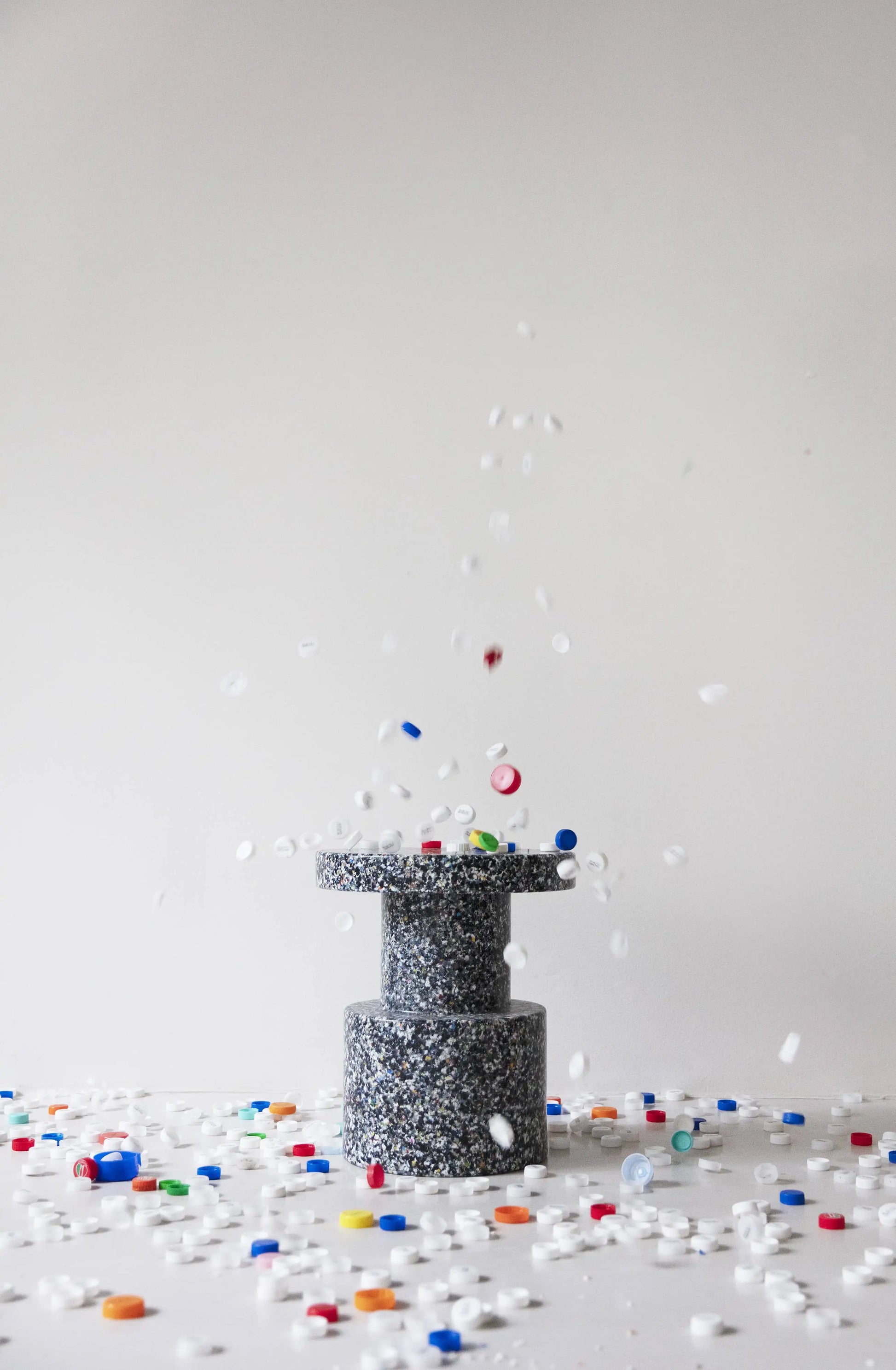 A cylindrical Normann Copenhagen Bit Stool sits on a white surface, surrounded by colorful plastic bottle caps. Some caps float mid-air and others rest on top, showcasing the playful, multifunctional design by Normann Copenhagen.