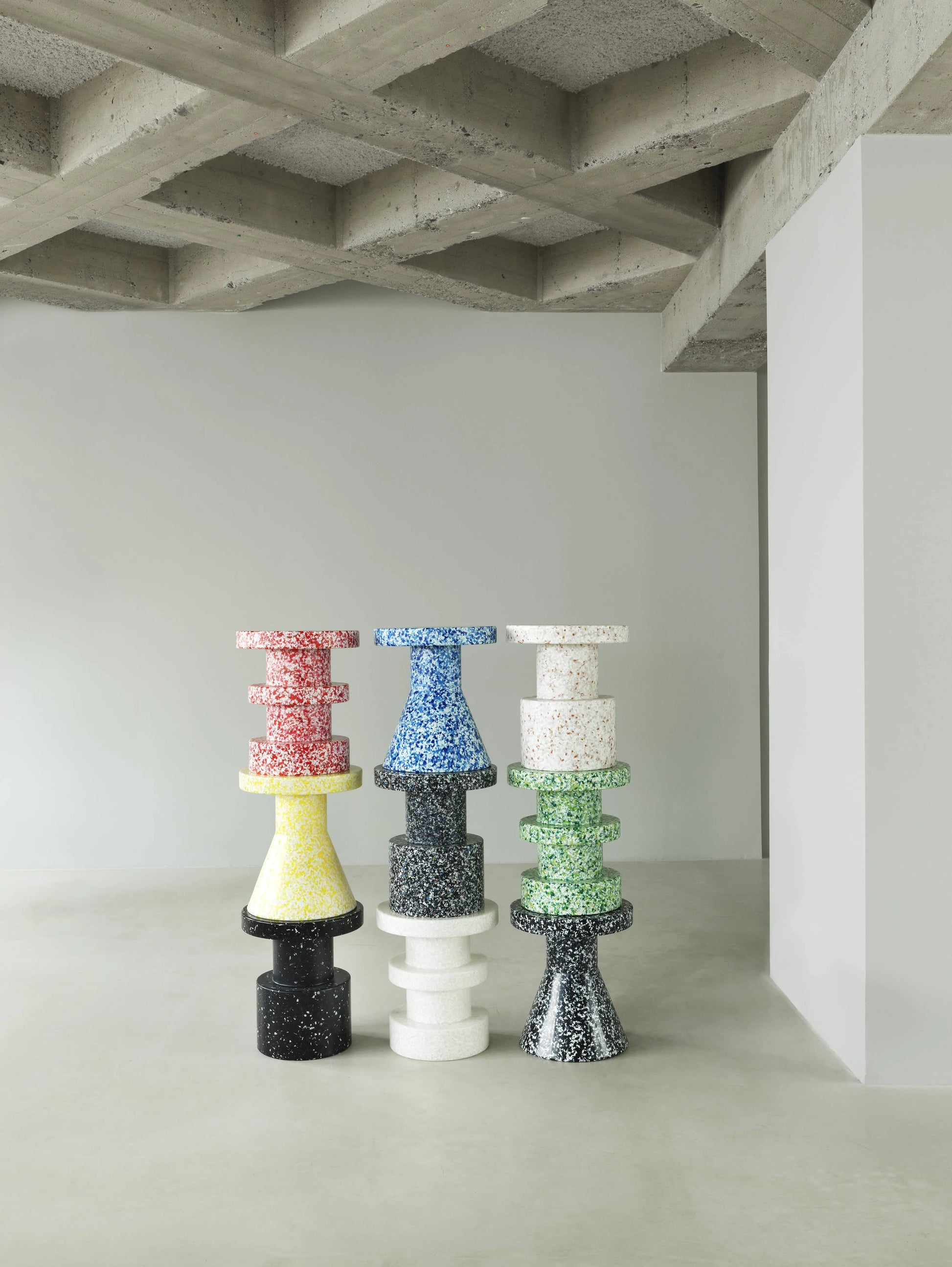 Six vibrant Normann Copenhagen Bit Stools are stacked in pairs against a minimalist white room with a concrete ceiling, each showcasing multifunctional design and bold terrazzo-patterned surfaces.