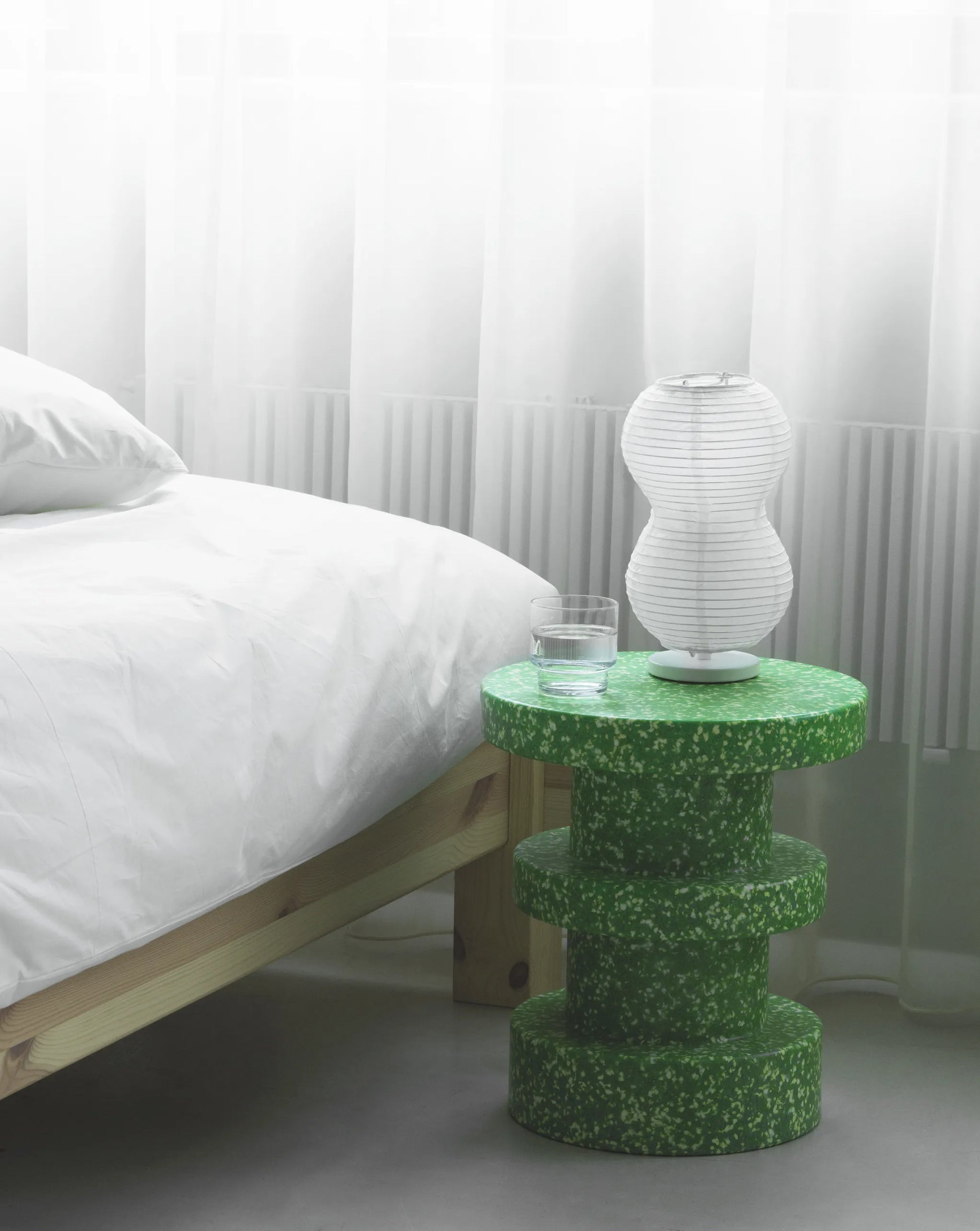 A minimal bedroom scene with a bed in white bedding, a green Normann Copenhagen Bit Stool Stack as a side table, a glass of water, and a sculptural white lamp, set before sheer curtains and soft natural light.
