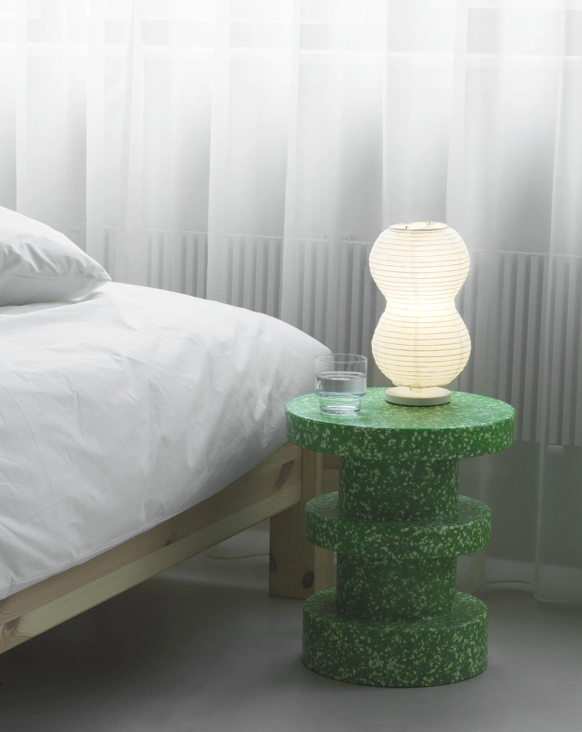 A modern bedside scene with a bed, a green Normann Copenhagen Bit Stool Stack as the side table, a glass of water, and a glowing lamp set against sheer white curtains.