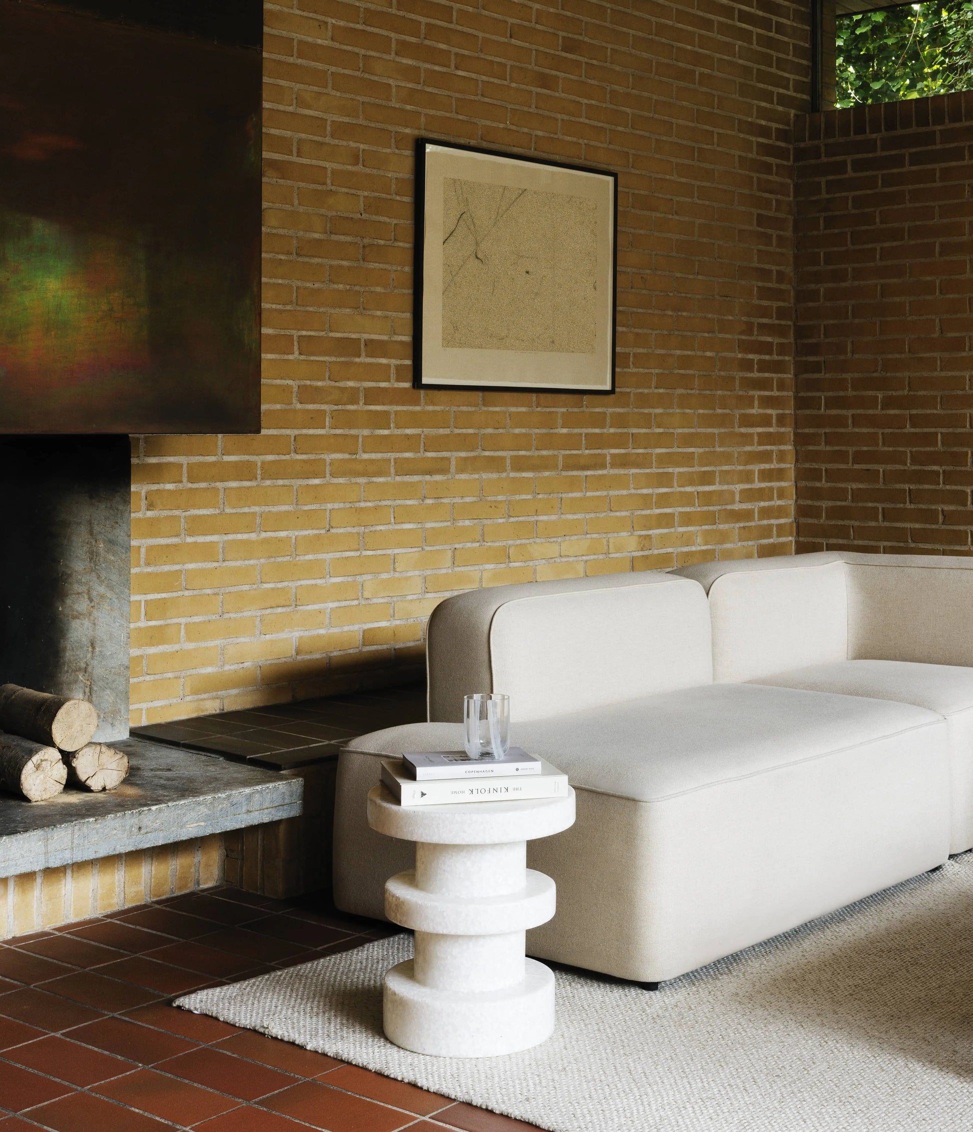 A modern living room with beige brick walls, a white sectional sofa, the Normann Copenhagen Bit Stool Stack as a stylish round side table with books and glasses, a fireplace with stacked logs, and framed wall art.