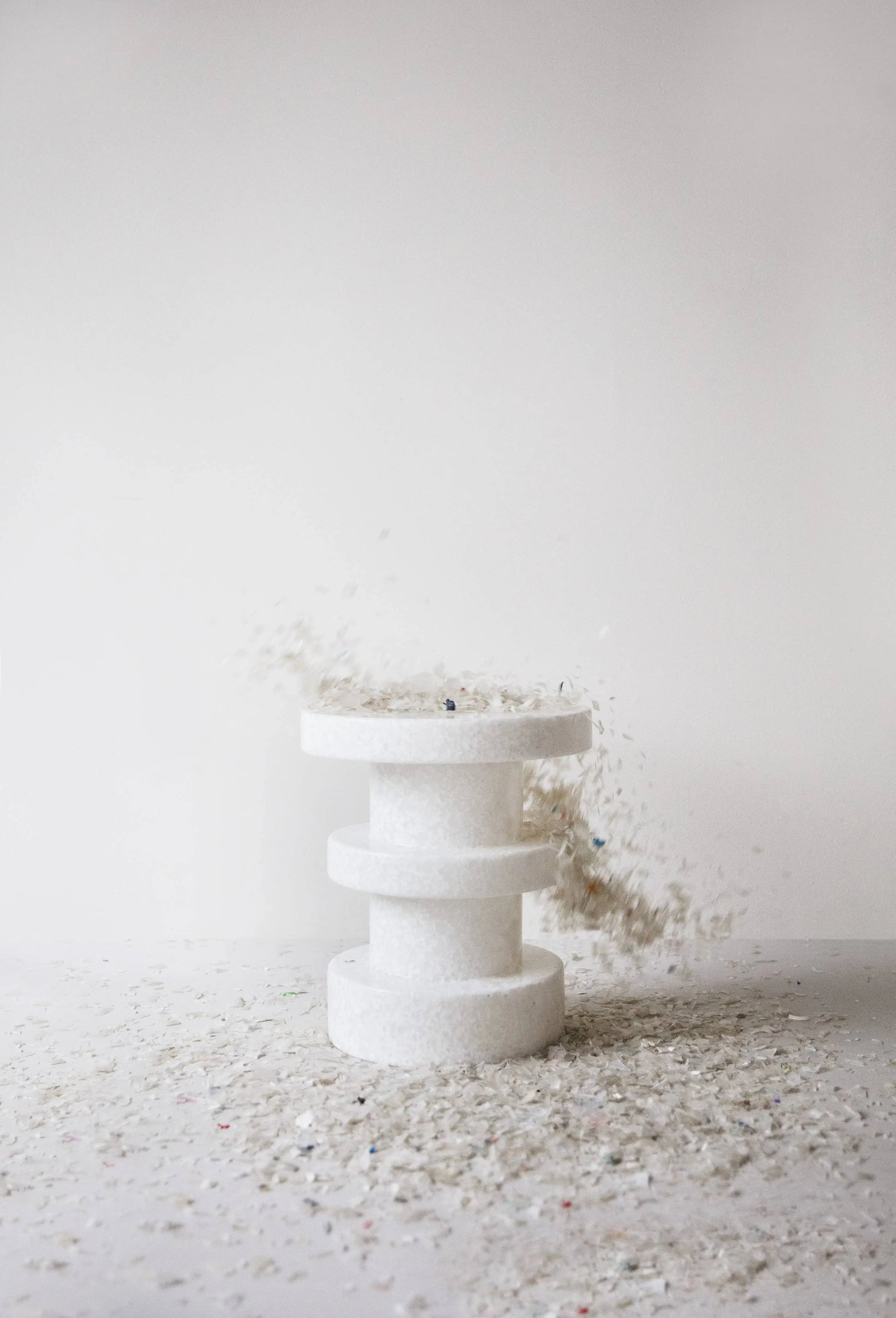 A modern, cylindrical white Normann Copenhagen Bit Stool Stack stands at the center of a minimalist scene, surrounded by scattered light-colored debris, with some pieces appearing mid-air beside it.