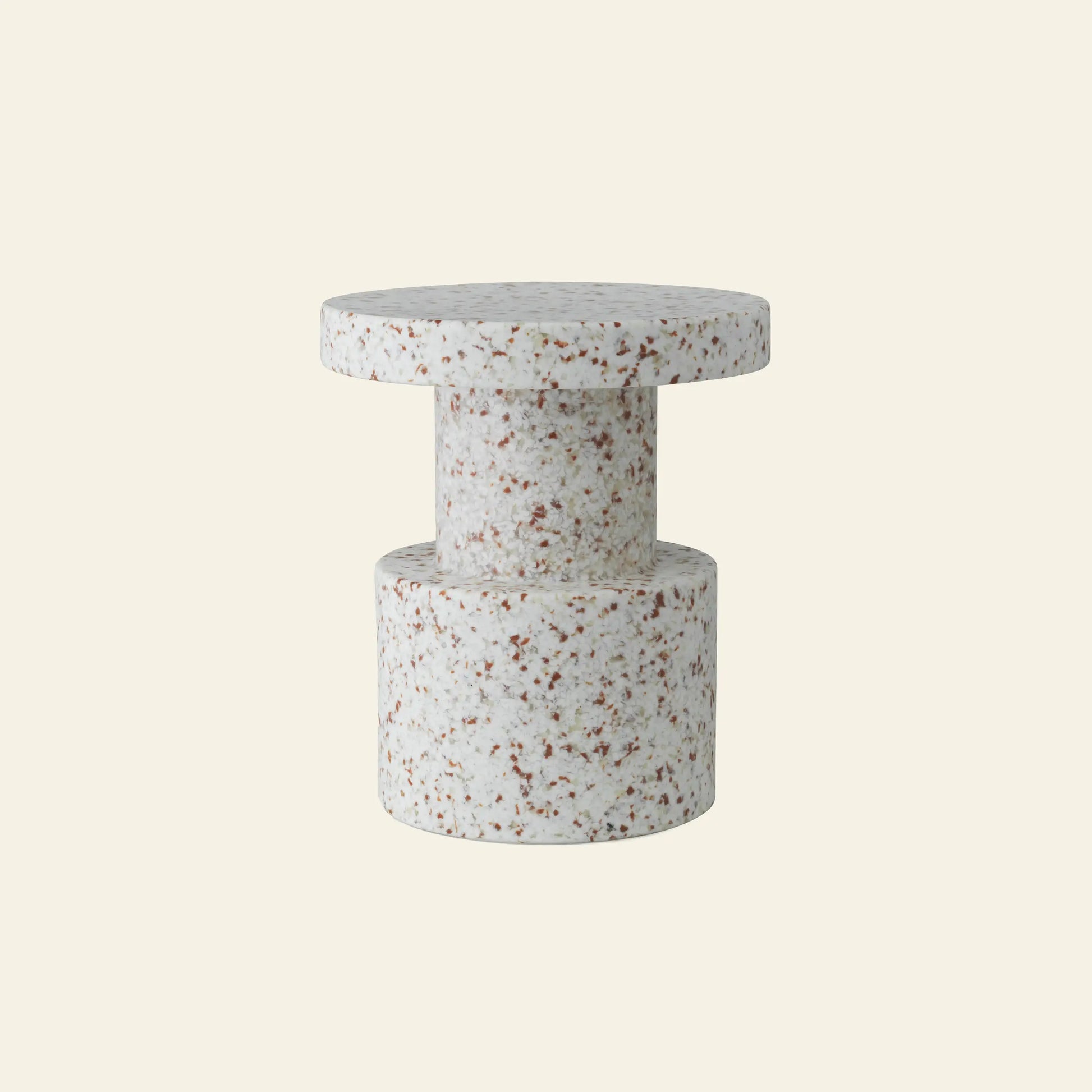The Normann Copenhagen Bit Stool by Normann Copenhagen is a modern cylindrical side table in speckled white and brown terrazzo, with a multifunctional design featuring a thick round top and wide base, shown against a plain beige background.