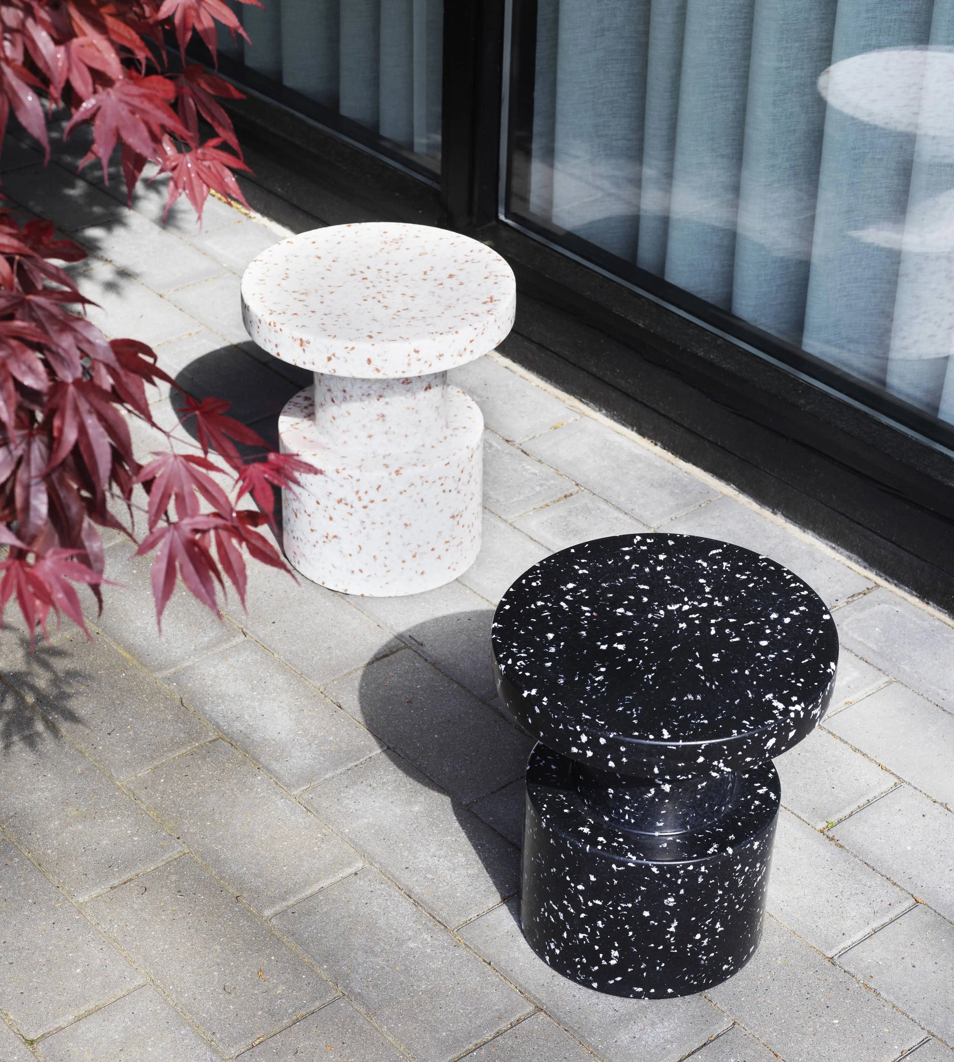 Two Normann Copenhagen Bit Stools—one white with reddish speckles, one black with white speckles—sit on a gray tiled patio beside a window and a leafy red plant.