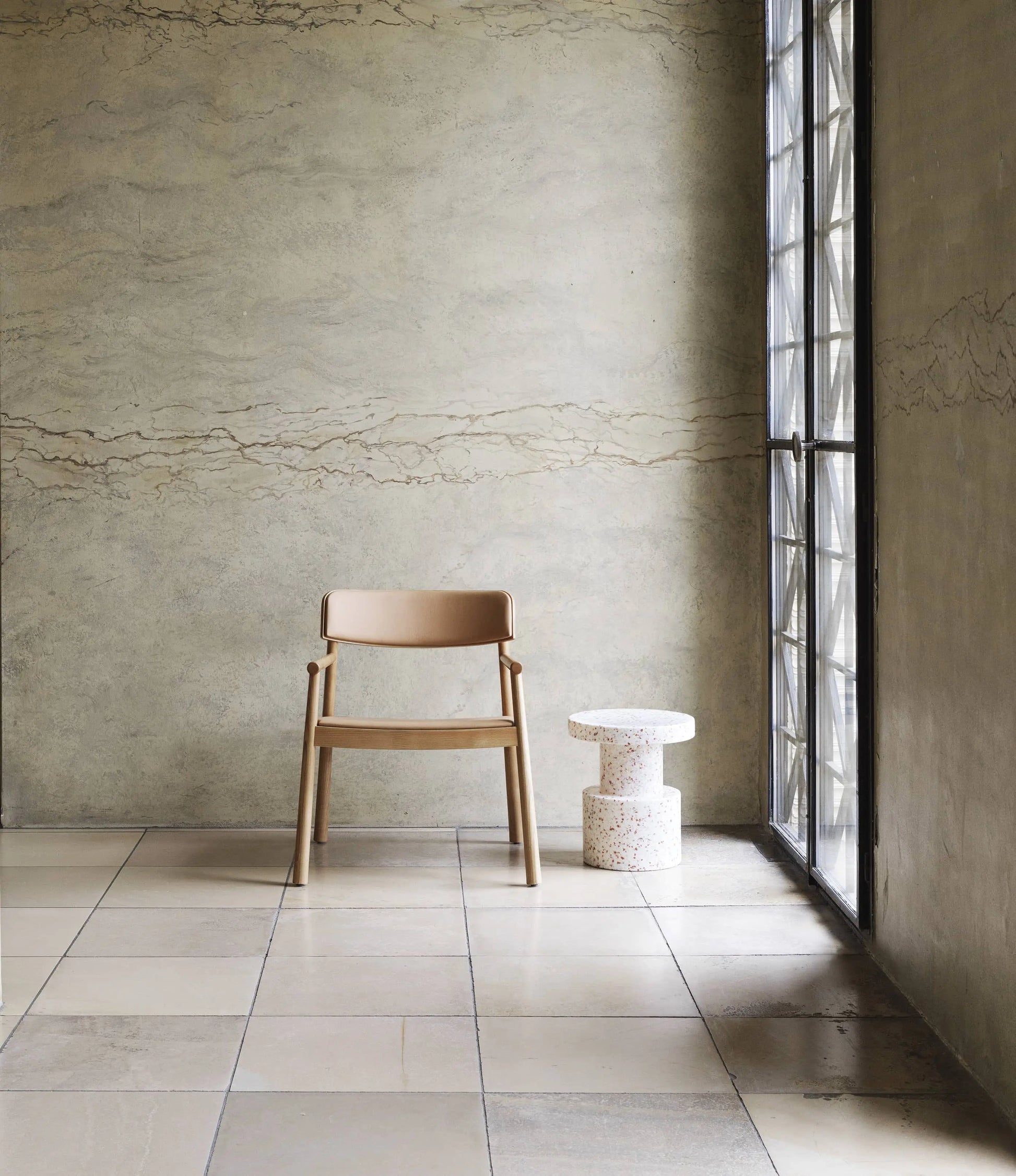 A wooden chair with a tan seat and backrest sits beside a Normann Copenhagen Bit Stool near a large window in a minimalist room with tiled floors and a textured beige wall, highlighting versatile seating by Normann Copenhagen.