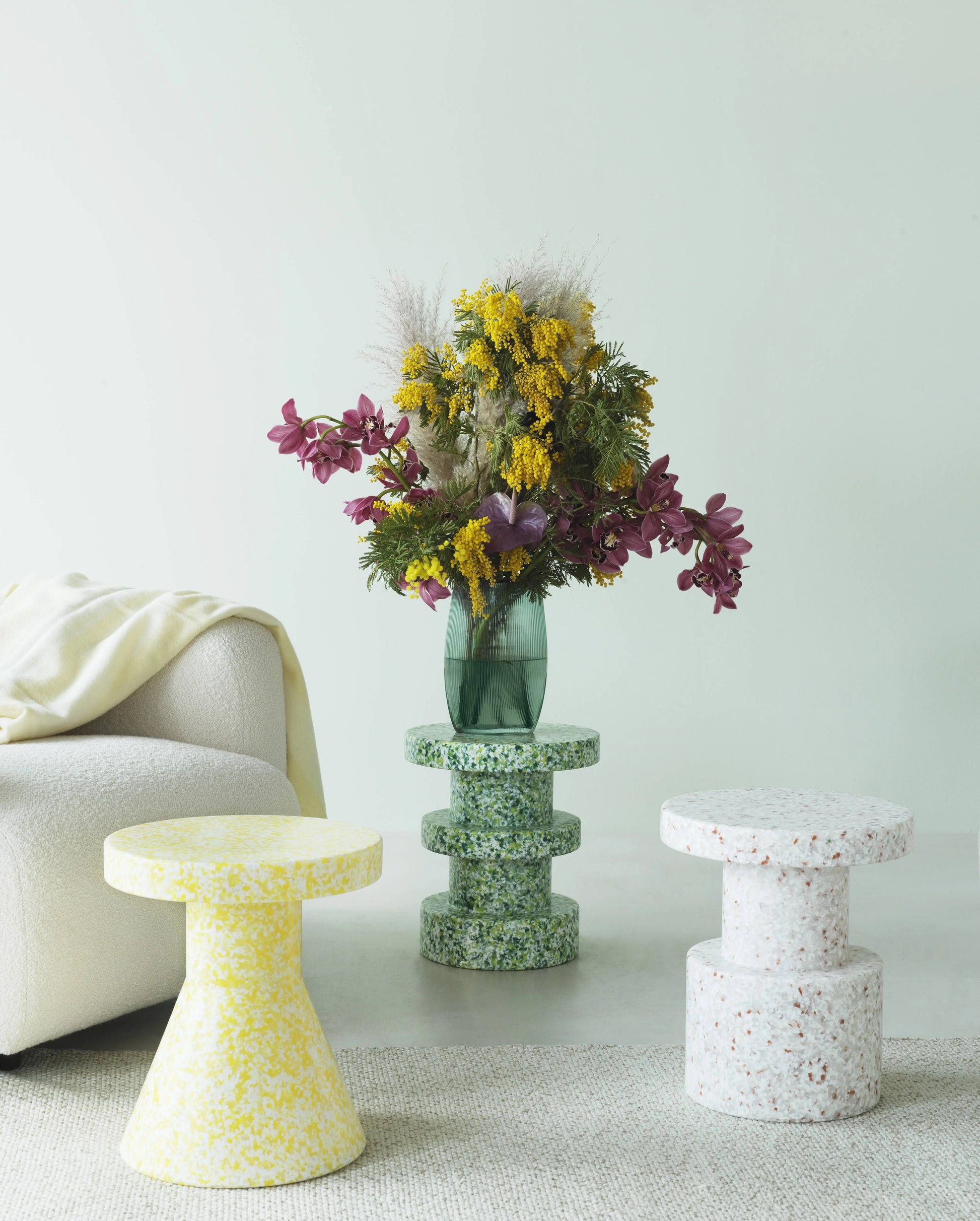A modern living space displays a white textured chair, cream blanket, and three Normann Copenhagen Bit Stools in yellow, green, and white as side tables, plus a green vase with vivid yellow and purple flowers—highlighting versatile design.