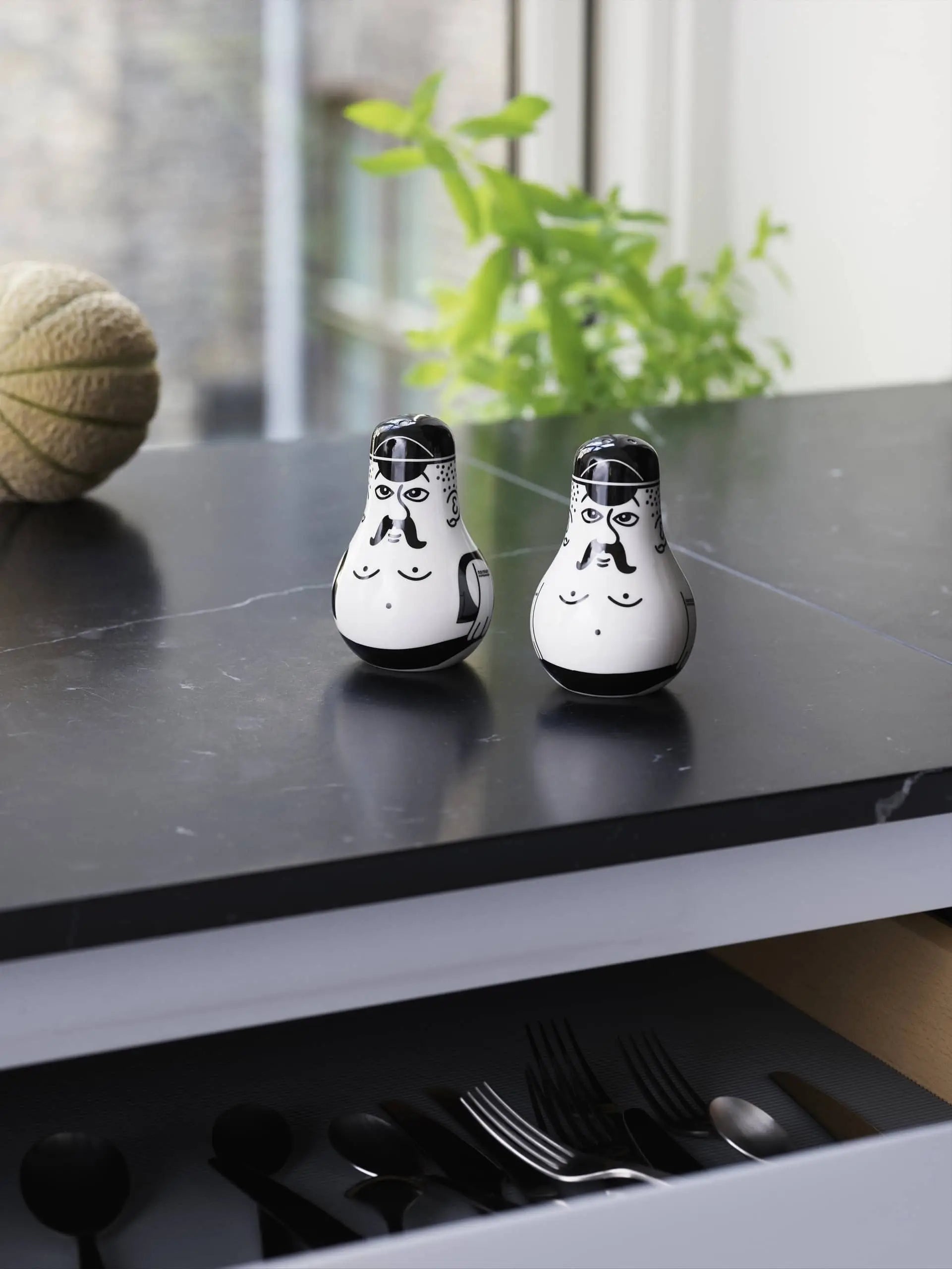 The Normann Copenhagen Friends Salt & Pepper Set features two ceramic shakers with painted faces and moustaches on a black countertop, complemented by a green plant and twine—an eye-catching, unique kitchen gift from Normann Copenhagen.