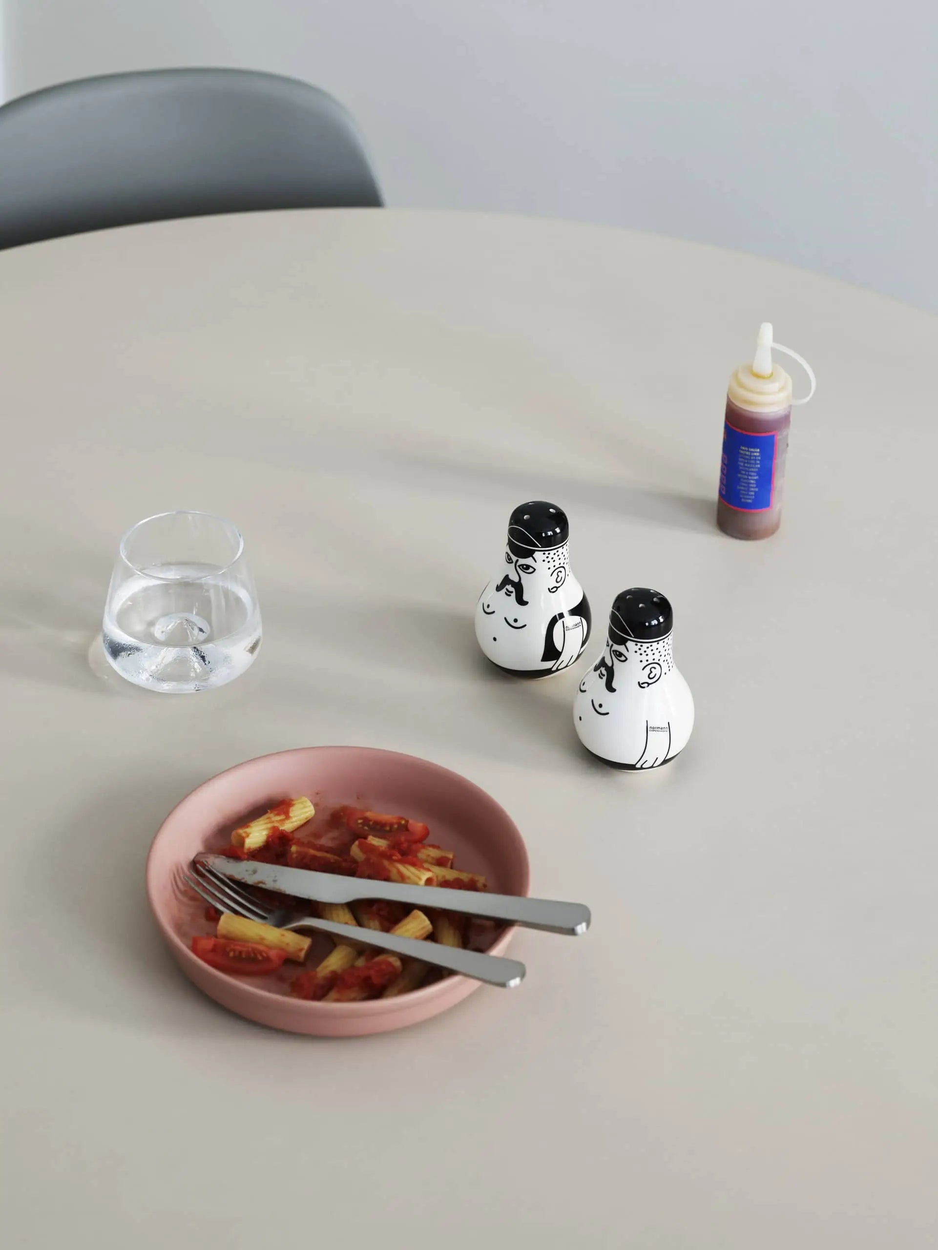 A round table set with a pink bowl of pasta, cutlery, water glass, sauce bottle, and the Normann Copenhagen Friends Salt & Pepper Set—an illustrated piece by Normann Copenhagen that makes a unique gift.