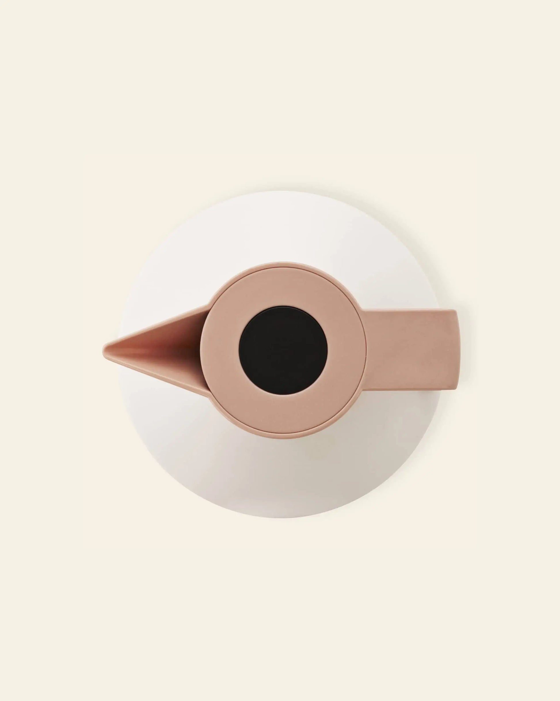 A top-down view of the Normann Copenhagen Geo Vacuum Jug in light pink, featuring a handle and spout, is displayed on a white round plate against a light beige background.