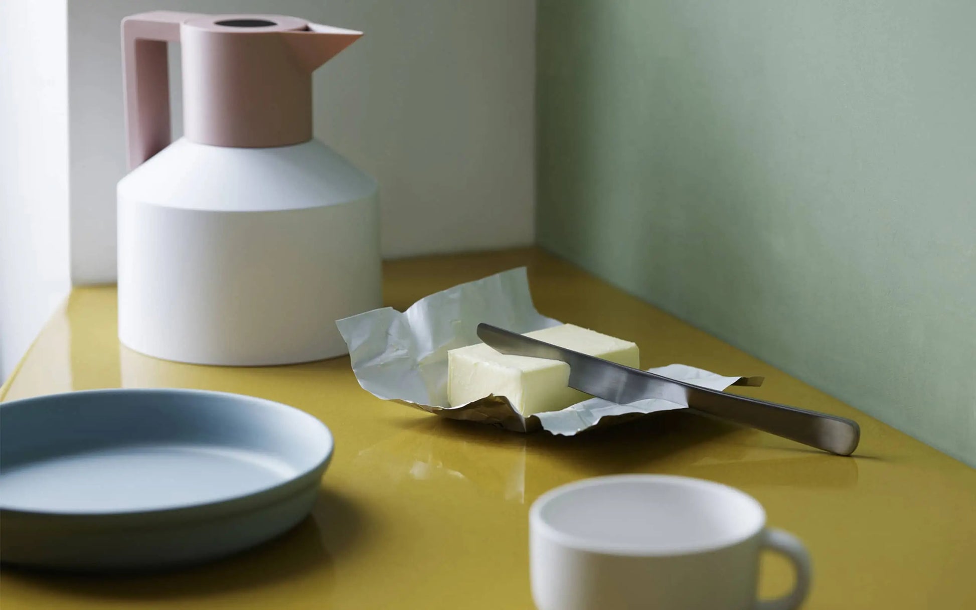 A butter knife rests on an opened stick of butter next to a blue plate, white cup, and the Normann Copenhagen Geo Vacuum Jug by Normann Copenhagen on a yellow countertop with a green wall in the background.
