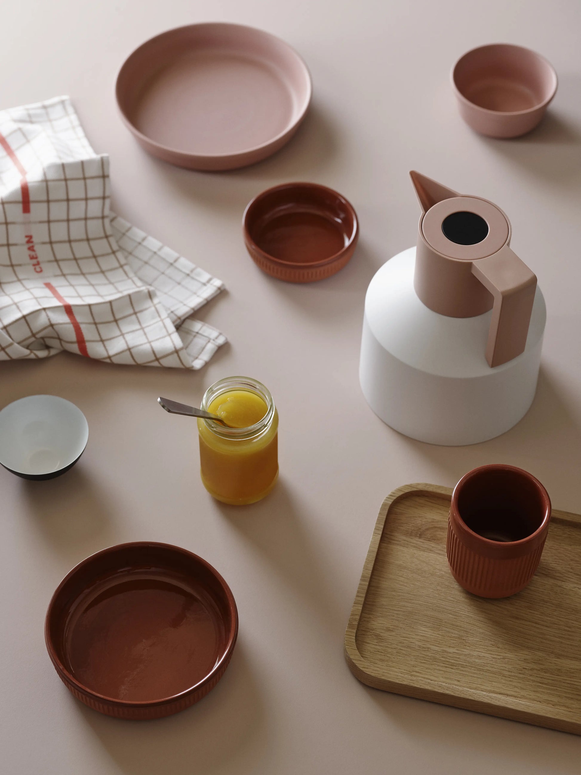 A minimalist breakfast setup features pink and brown ceramics, a Normann Copenhagen Geo Vacuum Jug with vacuum insulation, a jar of yellow spread with a spoon, a checkered cloth, and a wooden tray on a pale surface.