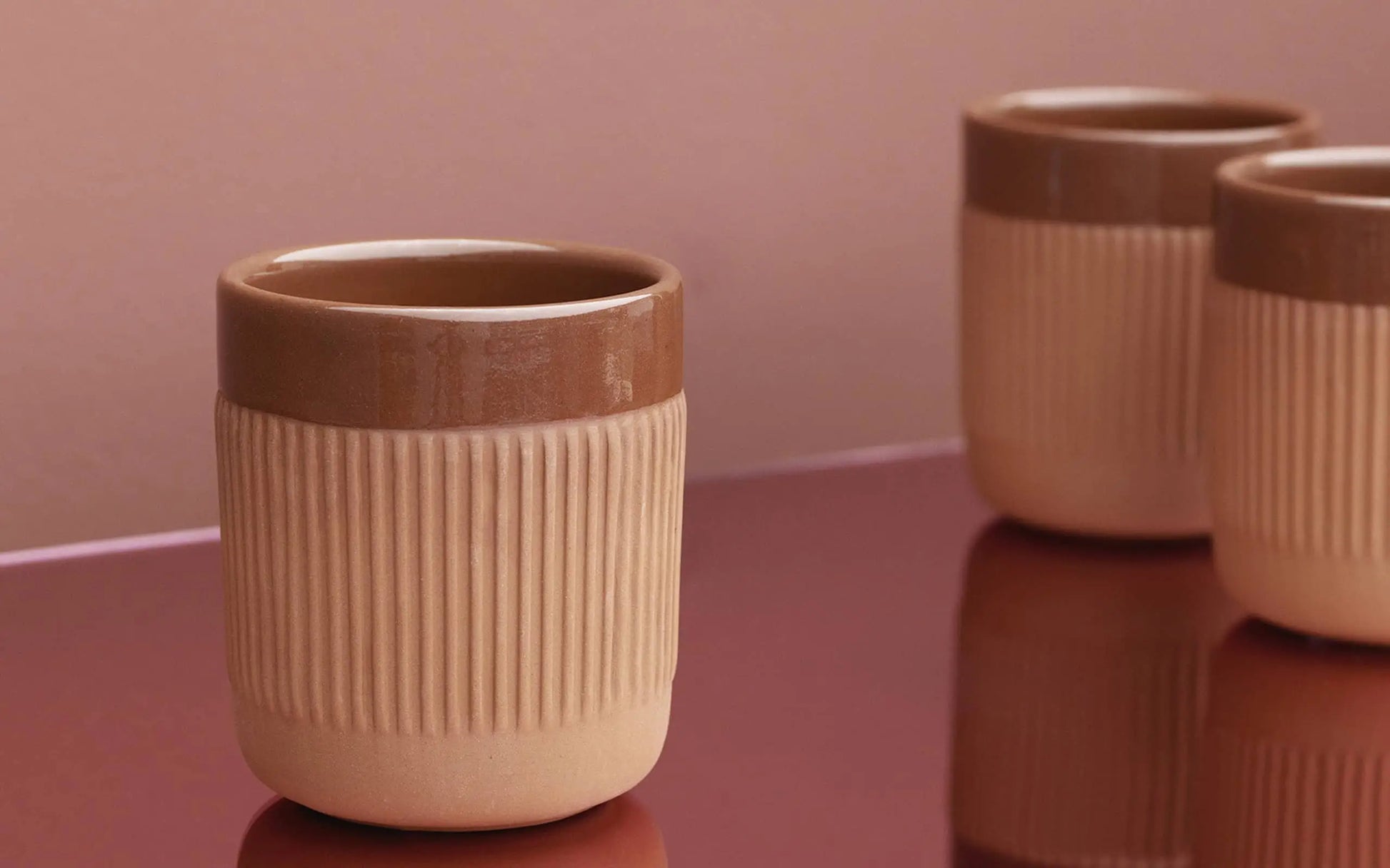 Three Normann Copenhagen Junto Cups with ribbed beige bases and glossy brown rims rest on a reflective, terracotta-hued surface against a soft pink background, with the foreground cup inviting you to enjoy a cosy night in.