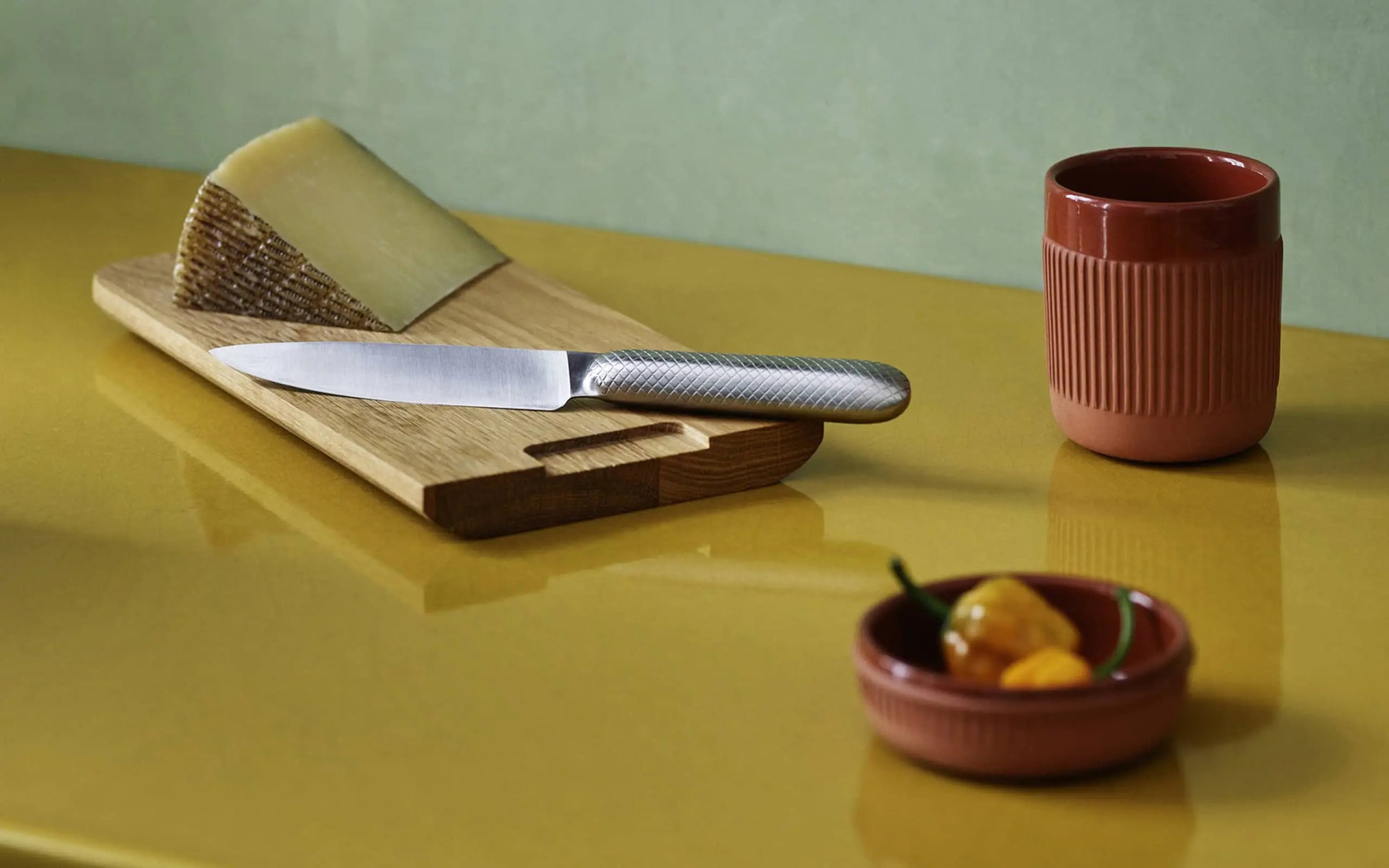 A metal knife on a wooden board with cheese sits beside the Normann Copenhagen Junto Cup in terracotta and a small dish of yellow and green peppers, all arranged on a glossy yellow surface—ideal for a cosy night in.