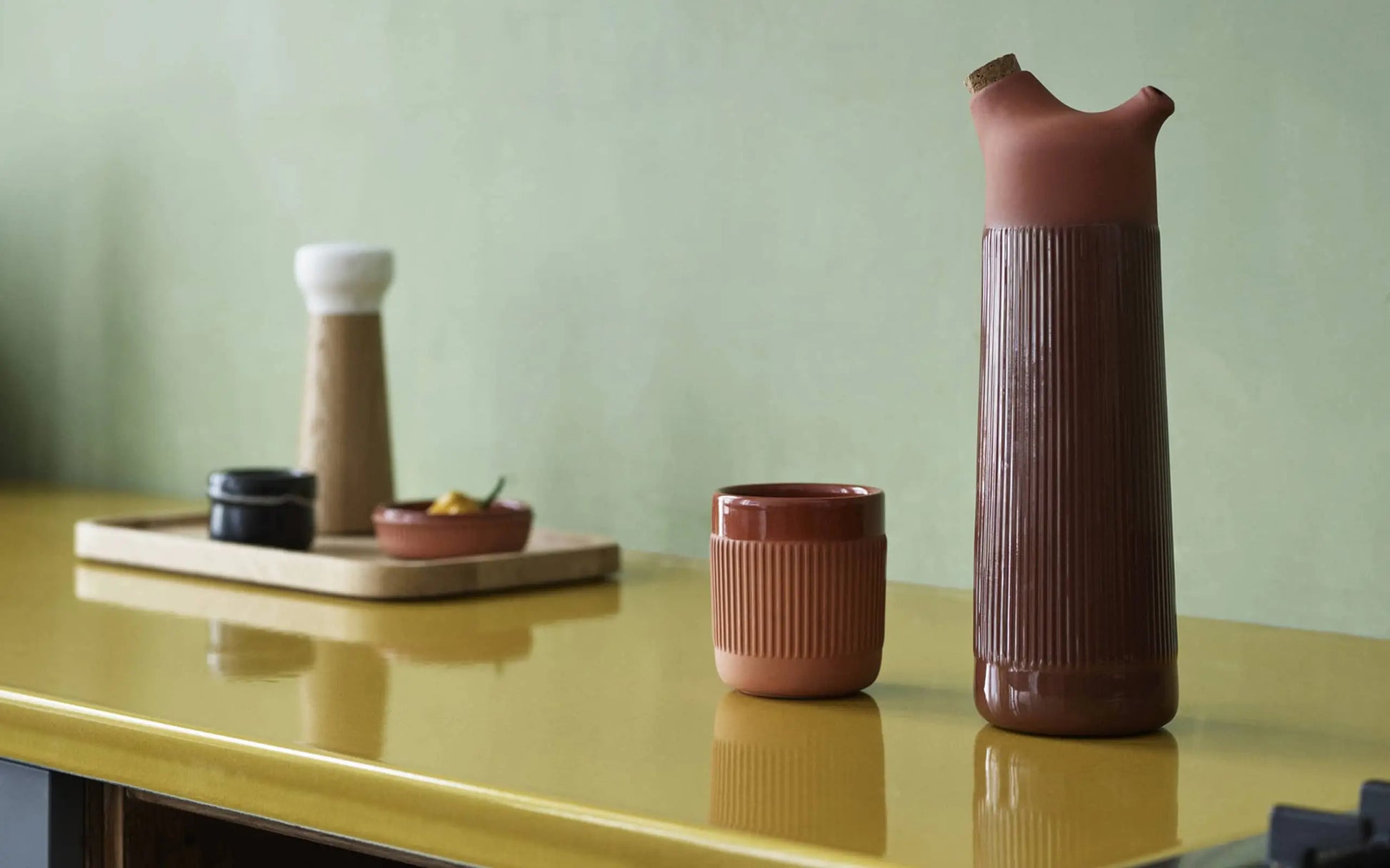 A tall, ribbed ceramic jug and the Normann Copenhagen Junto Cup in terracotta rest on a glossy yellow counter. A wooden tray with kitchenware sits behind, set against a pale green wall—ideal for a cosy night in.