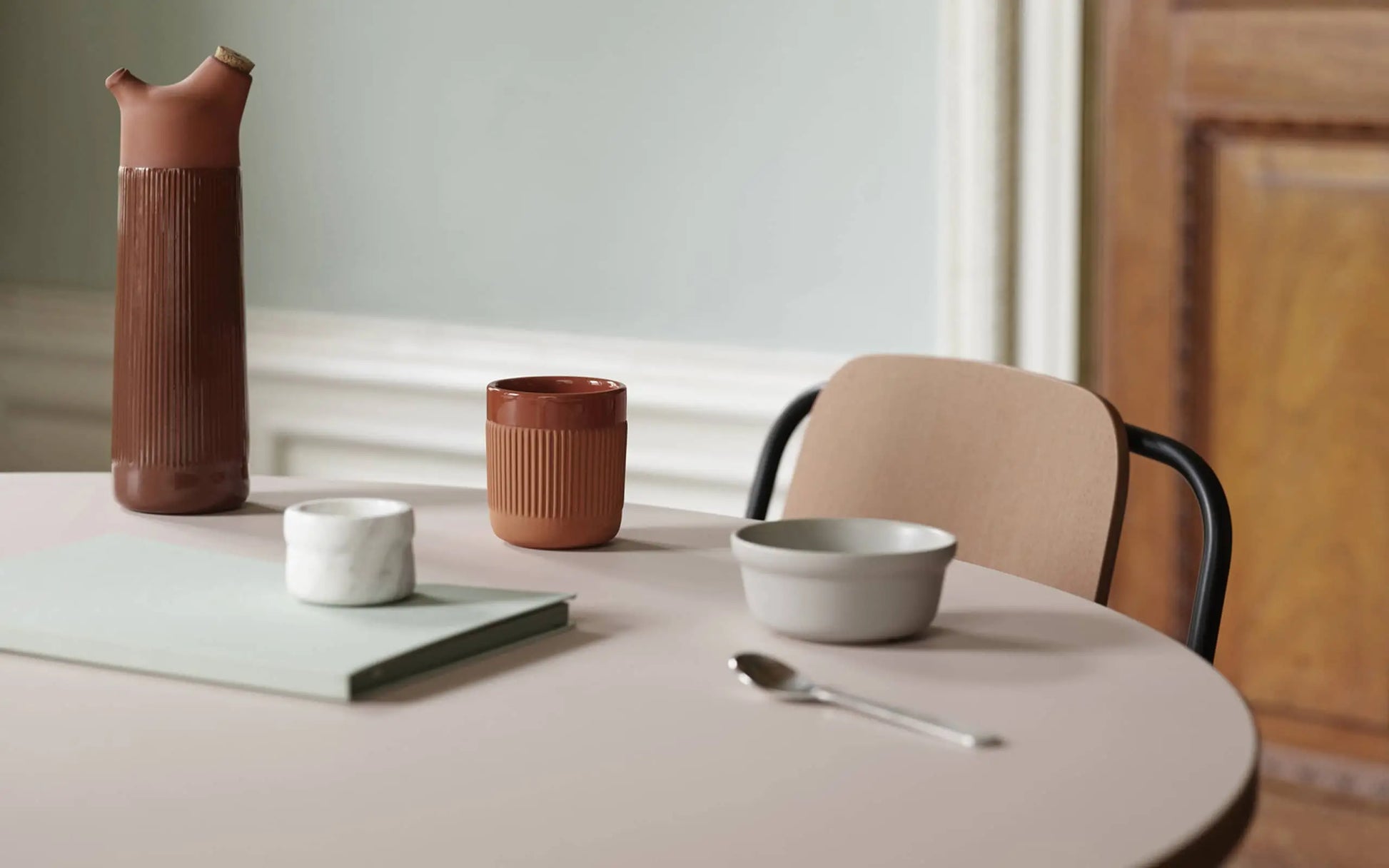 A minimalist table setting in terracotta features a brown pitcher, Normann Copenhagen Junto Cup, white bowl, white marble container, green notebook, and spoon on a light table by a beige chair—perfect for a cosy night in.