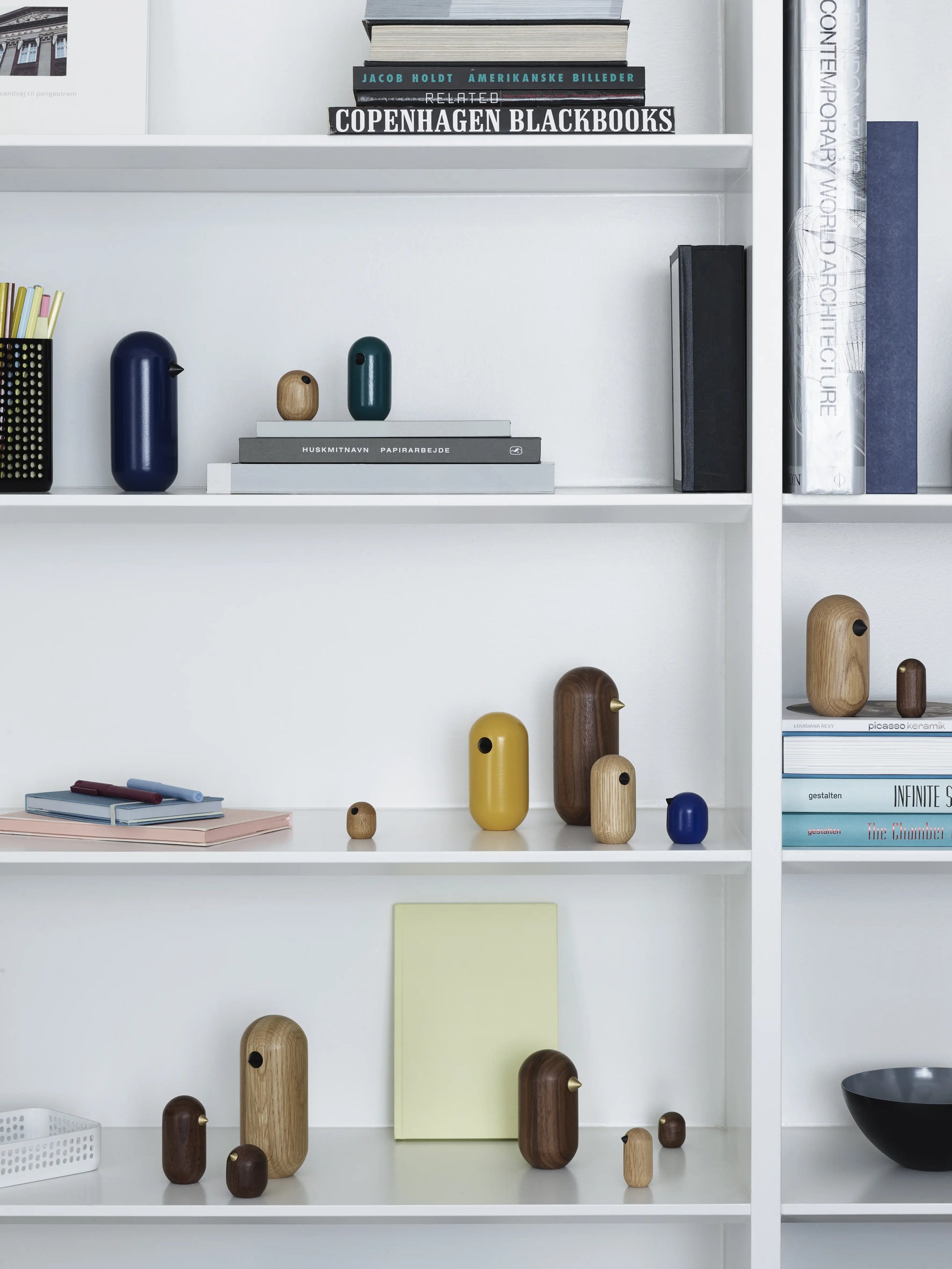 A white bookshelf holds books, stacked magazines, art prints, and several Normann Copenhagen Little Bird figurines (4.5cm) in brown, yellow, blue, and natural wood tones—each a unique decor piece by Normann Copenhagen.