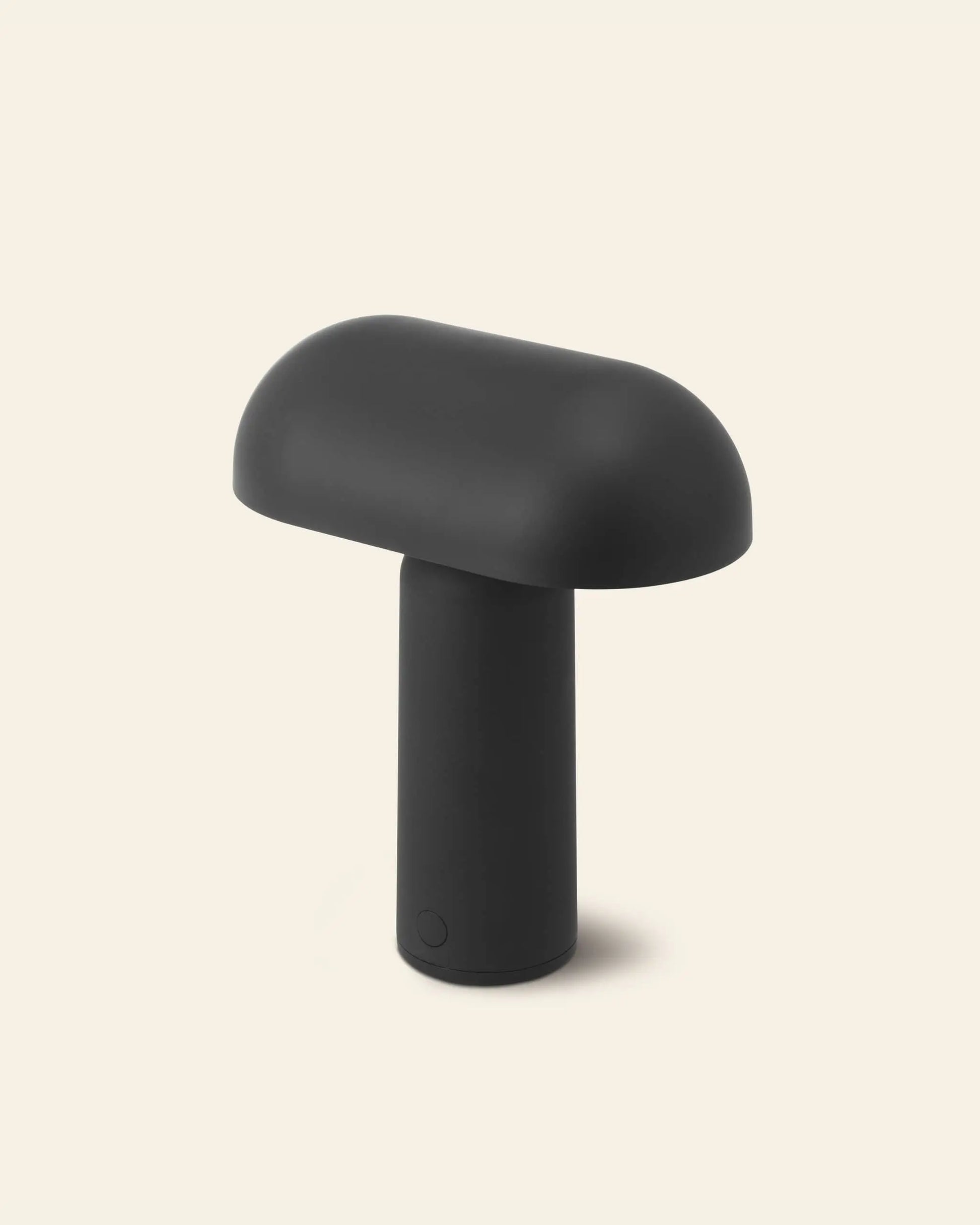 The Normann Copenhagen Porta Table Lamp features a modern, minimalist mushroom shape with a matte black finish and cylindrical base, capturing the brand’s sleek style. Set against a beige background, it stands out as a stylish portable lamp.