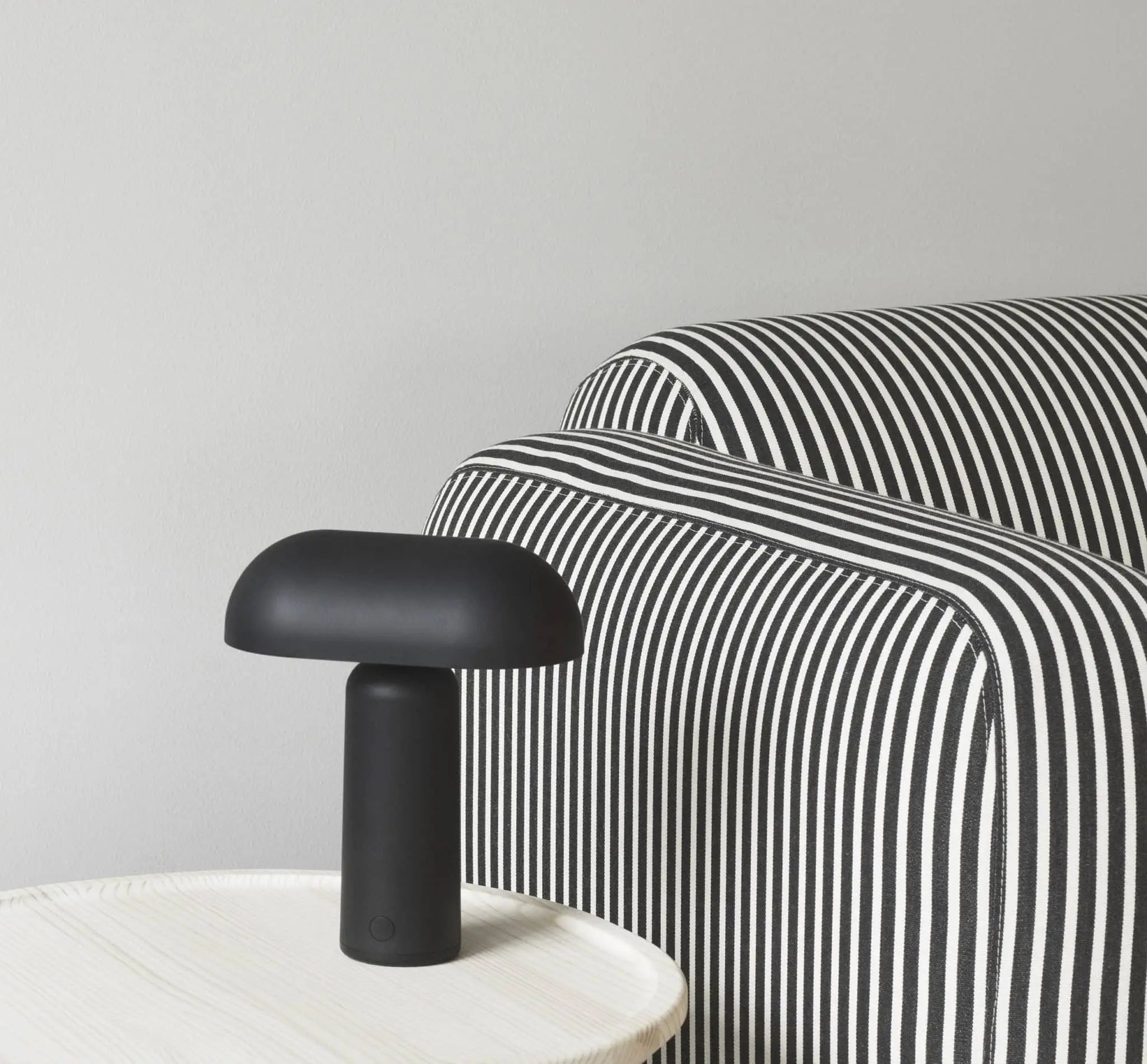 A Normann Copenhagen Porta Table Lamp with a modern design sits on a light-colored round table beside a sofa with black and white stripes, set against a plain light gray wall.