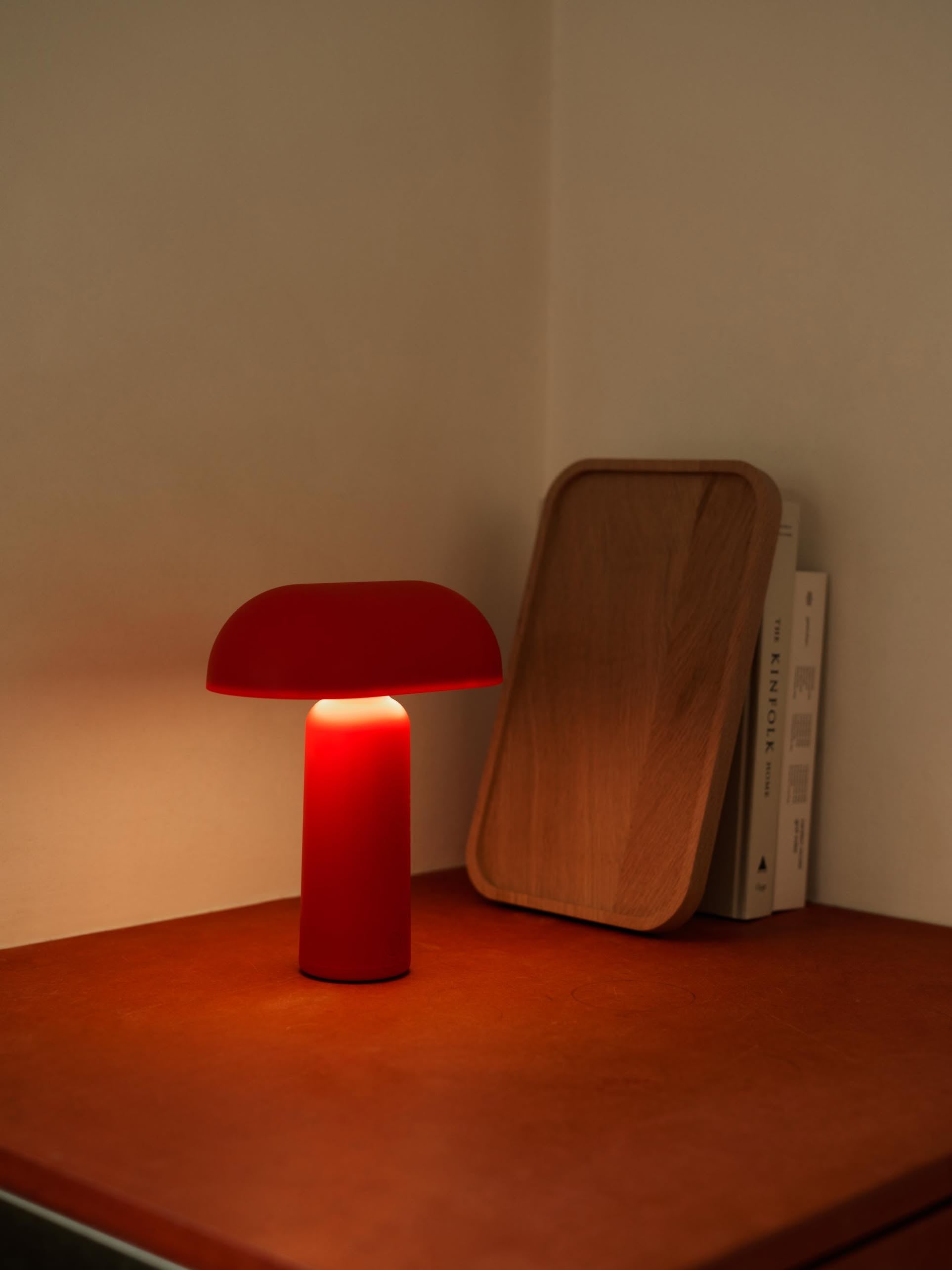 A Normann Copenhagen Porta Table Lamp in red glows on a reddish-brown surface beside an upright wooden tray and two books, creating a cozy, softly lit corner.