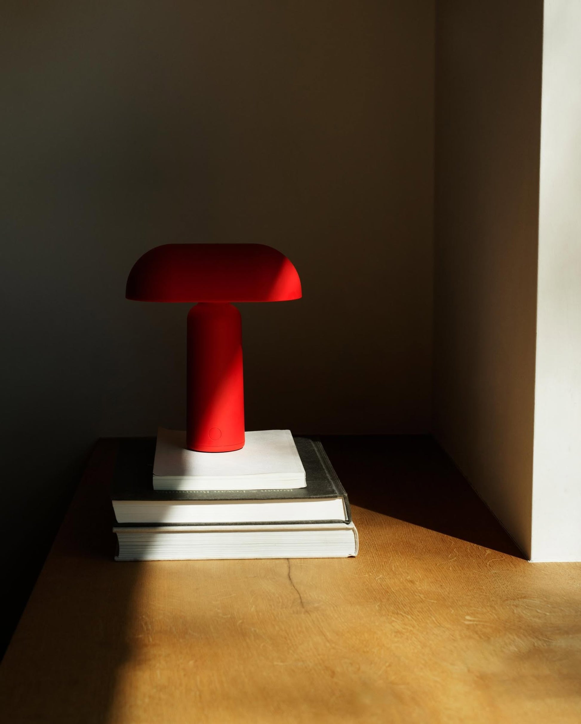 A Normann Copenhagen Porta Table Lamp in red rests on a stack of three books on a wooden surface, sunlight streaming through the window. The lamp’s modern design pops against a minimalist, neutral backdrop.