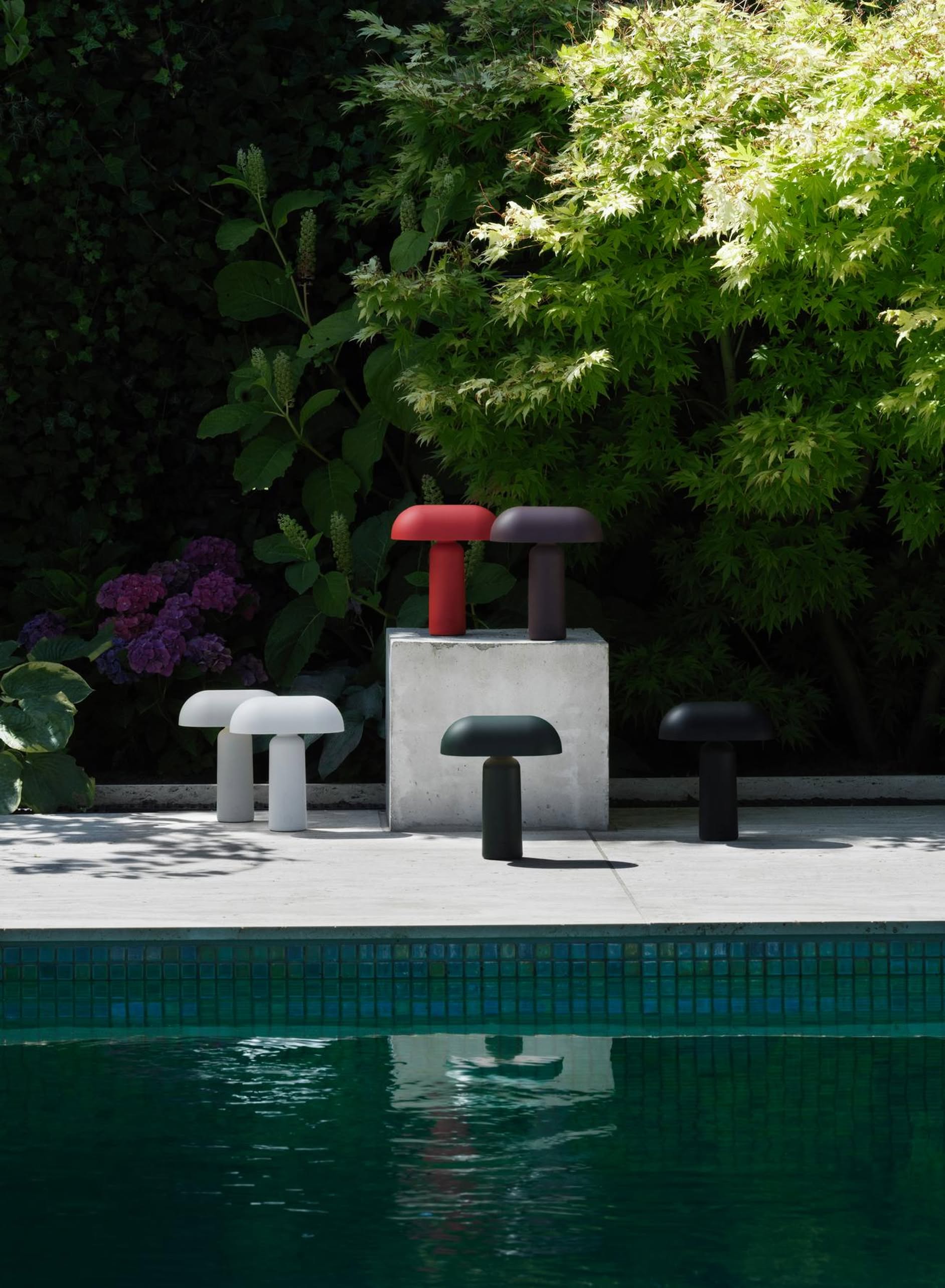 Five Normann Copenhagen Porta Table Lamps in white, red, black, and gray are displayed on a concrete platform by the pool, their sleek modern design standing out among lush greenery and purple flowers.
