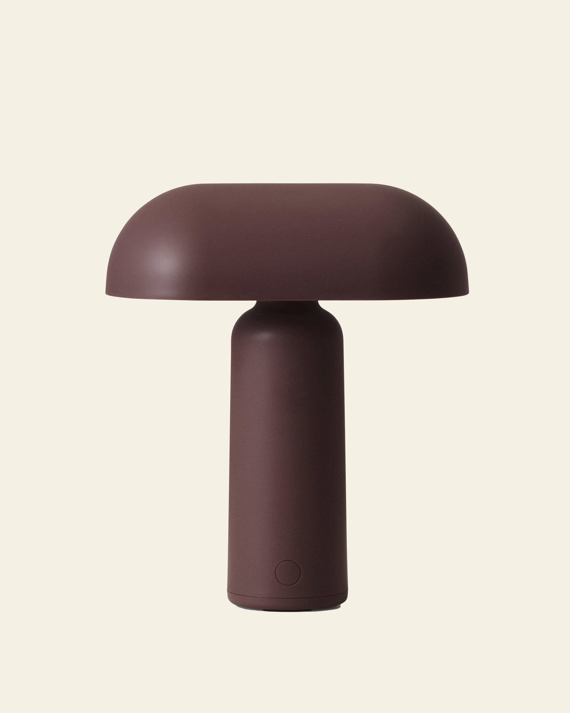 The Normann Copenhagen Porta Table Lamp features a minimalist design with a matte brown finish, cylindrical base, and wide dome-shaped shade, inspired by Modern Design. Shown against an off-white background.