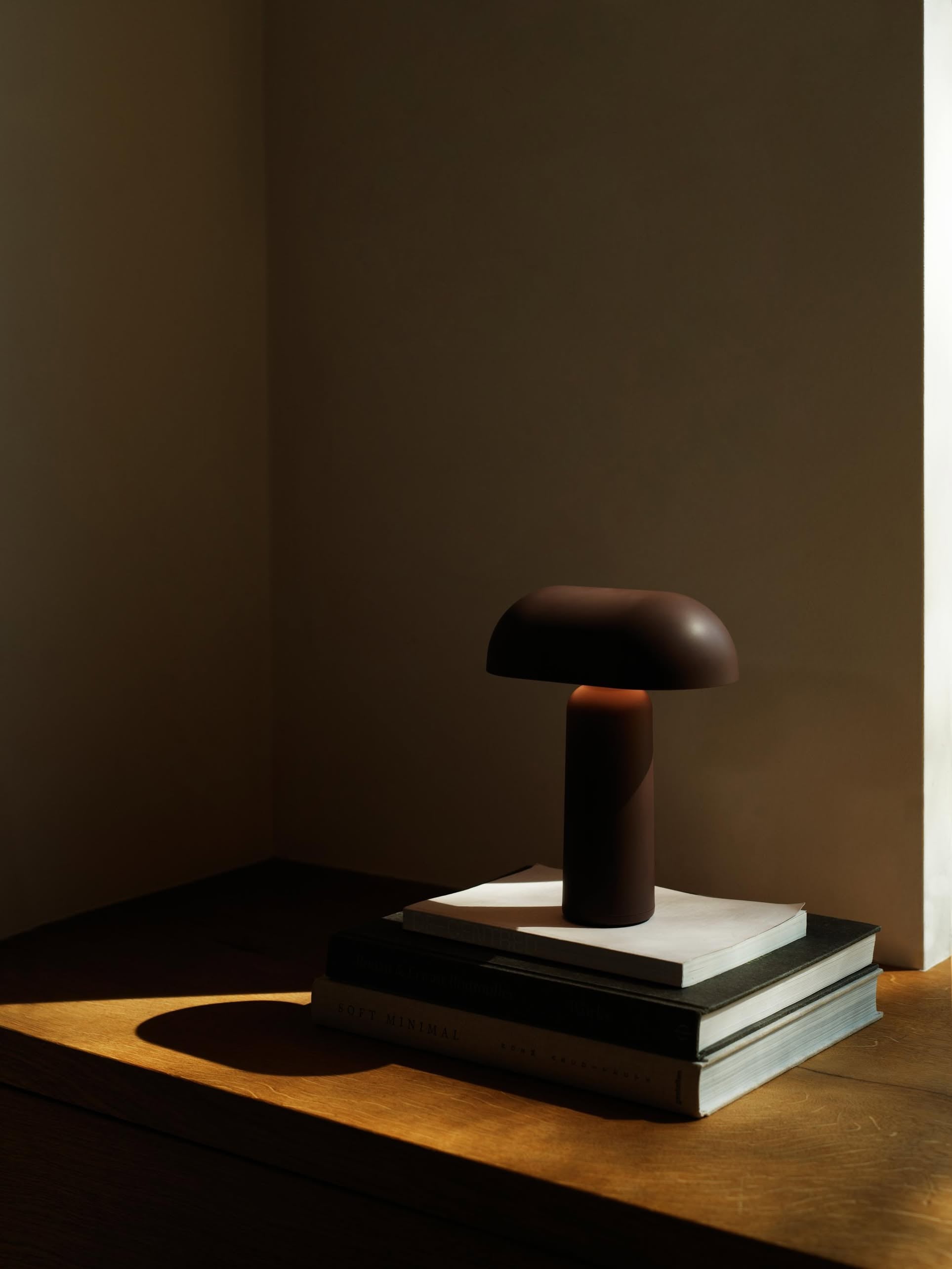 A compact Normann Copenhagen Porta Table Lamp sits atop stacked books in a sunlit corner, casting shadows on the wooden surface. The minimalist scene is illuminated by warm, natural light.