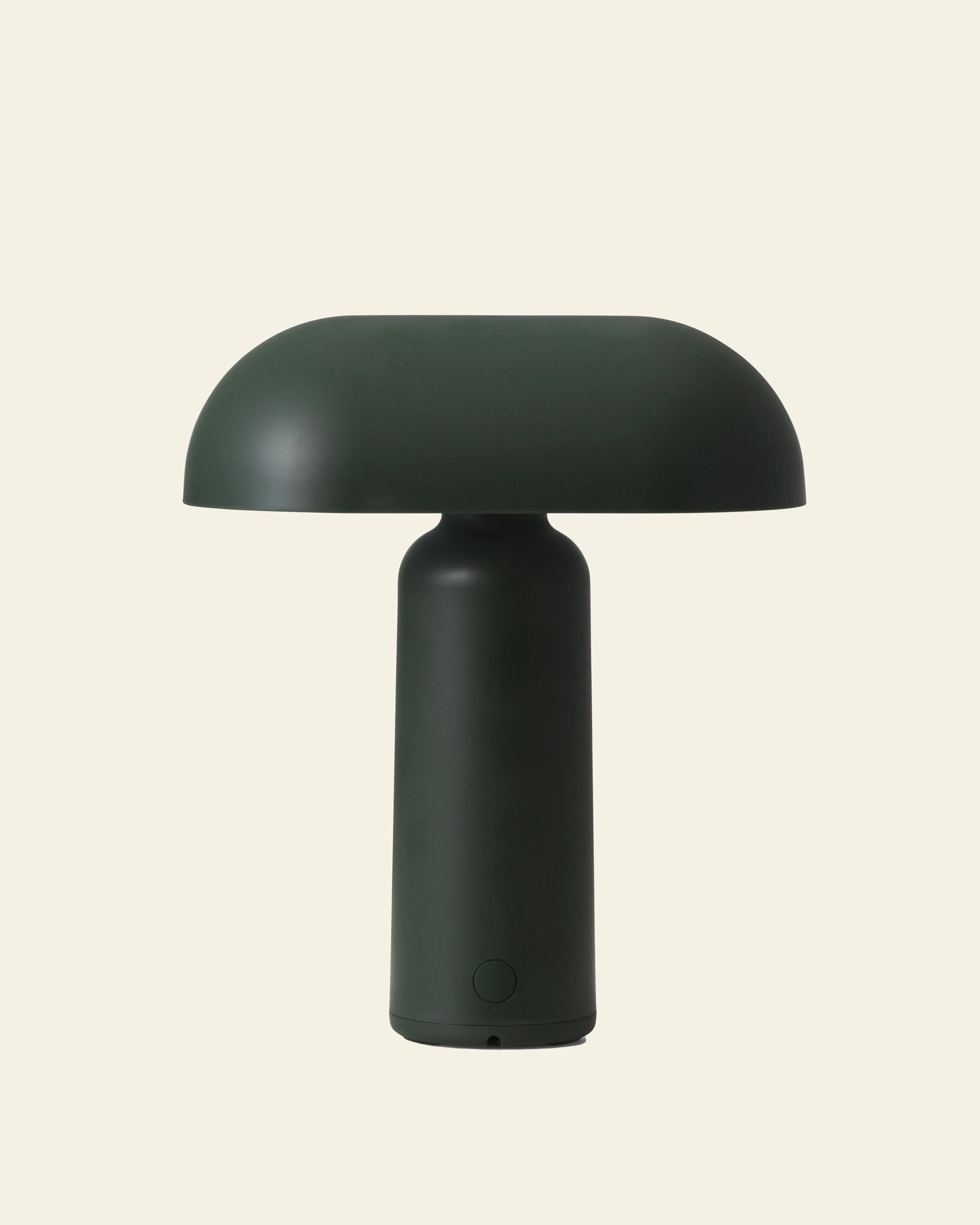 The Normann Copenhagen Porta Table Lamp features a modern, dark green mushroom shape with a rounded dome top and cylindrical base, inspired by Normann Copenhagen, and is shown against a plain beige background.