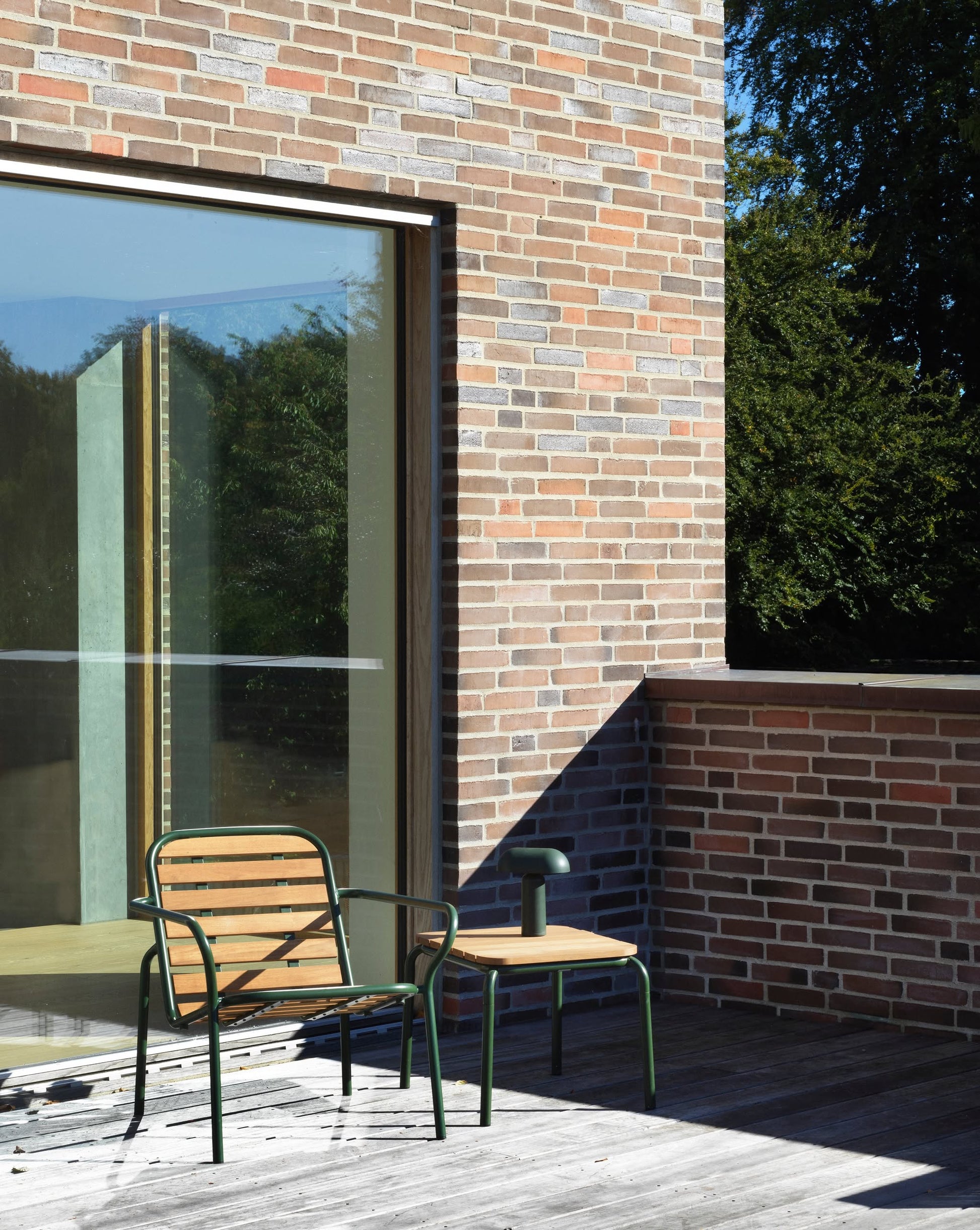 A modern patio showcases a wooden chair, small side table, and the Normann Copenhagen Porta Table Lamp on a wood deck beside a large glass door in a brick building, with greenery reflected in the glass.