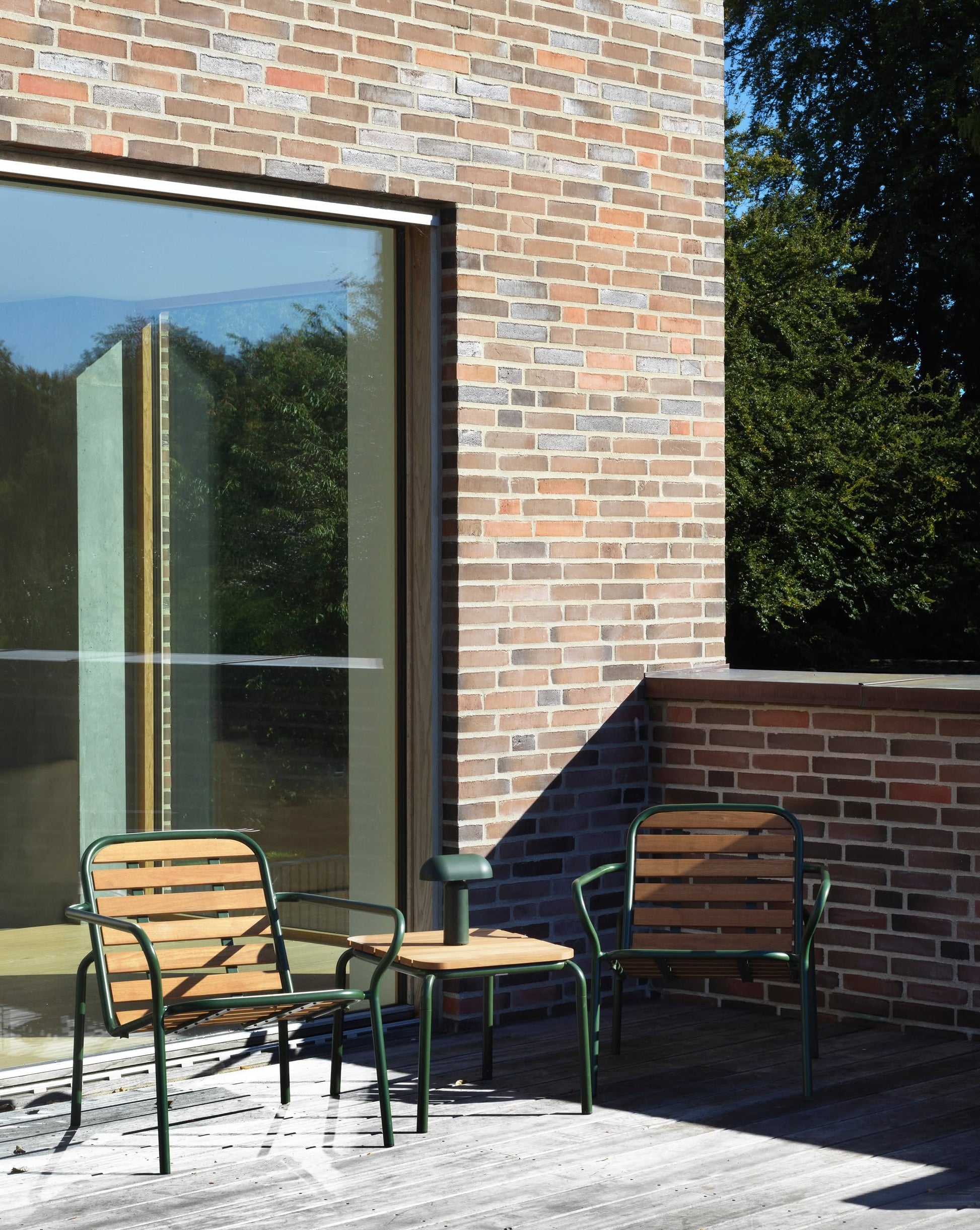 A Normann Copenhagen Porta Table Lamp sits on a small table between two metal chairs on a wooden deck outside a brick building with large windows reflecting trees and sunlight.