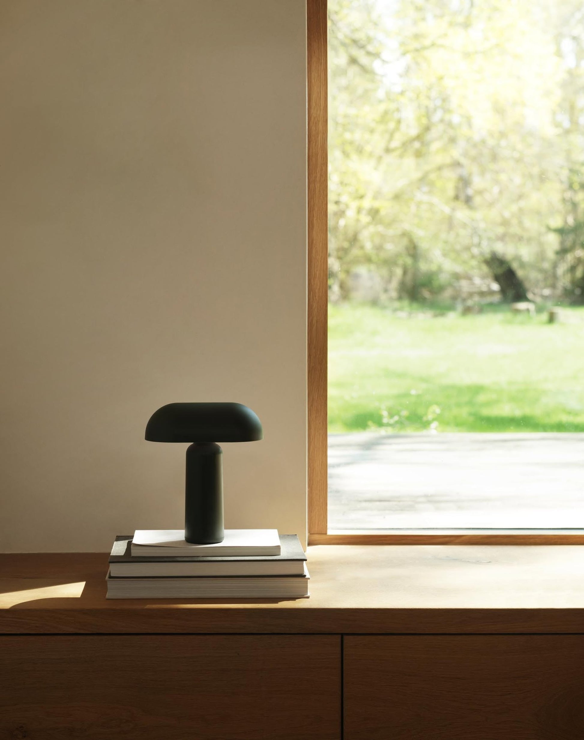 A sleek black Normann Copenhagen Porta Table Lamp rests on a stack of books by a large window, sunlight brightening the minimal space and offering a view of grass and trees outside.