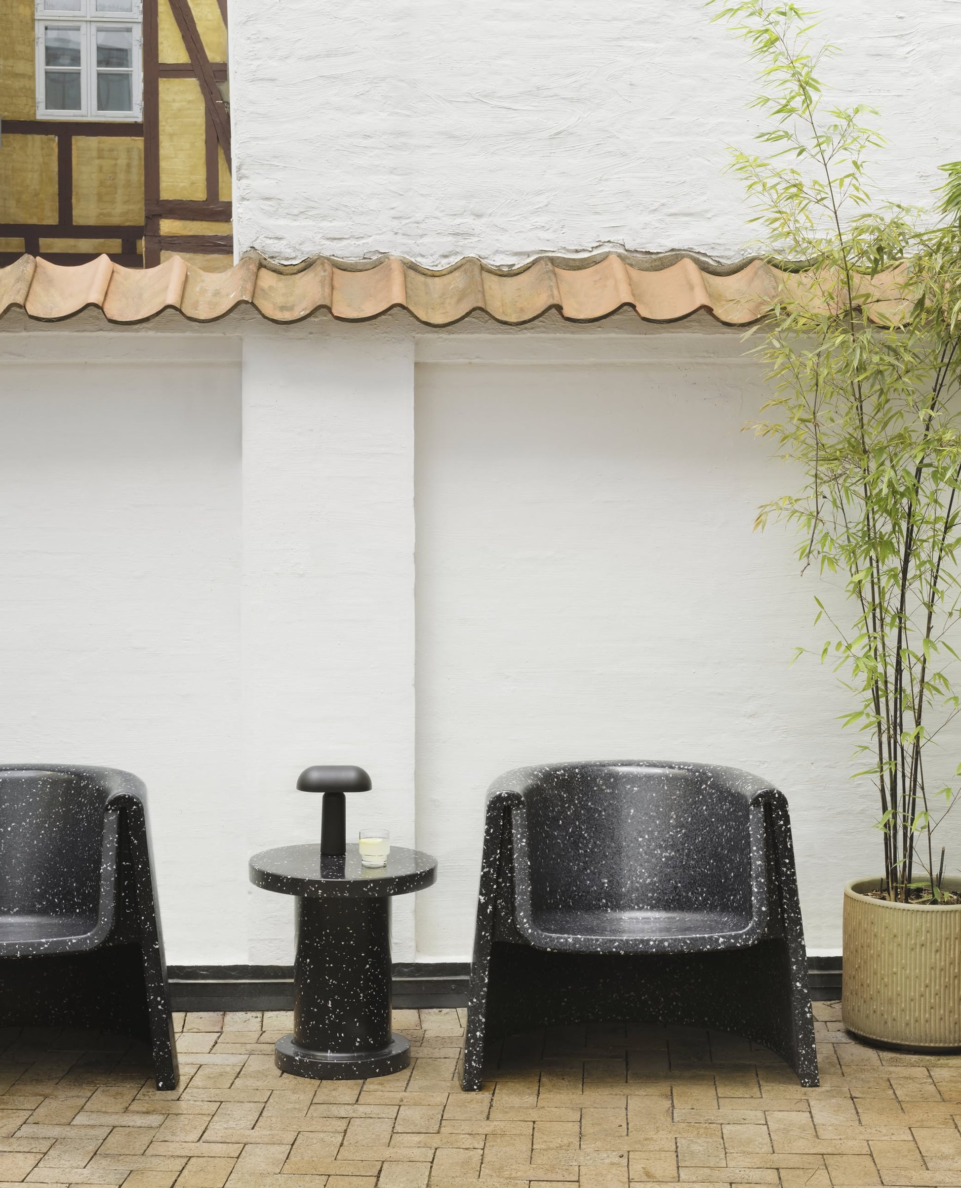 Two black speckled armchairs and a Normann Copenhagen Porta Table Lamp on a matching side table sit by a white wall with tiled roof. To the right is a tall potted bamboo, while brick paving covers the modern floor.
