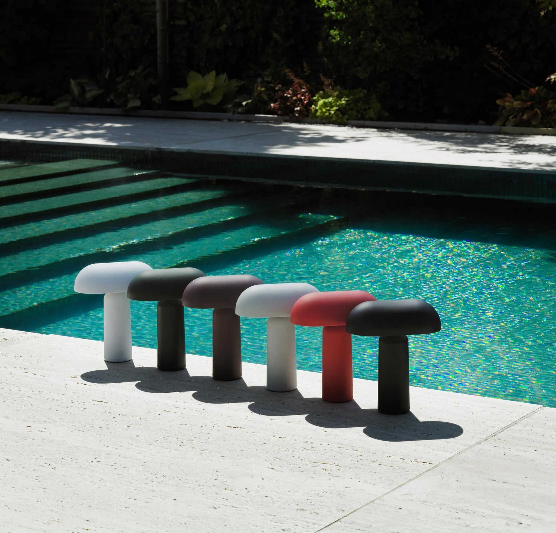 Six colorful Normann Copenhagen Porta Table Lamps from Normann Copenhagen are arranged on a light stone poolside, their modern design and portable style casting distinct shadows against the blue pool water and lush greenery.
