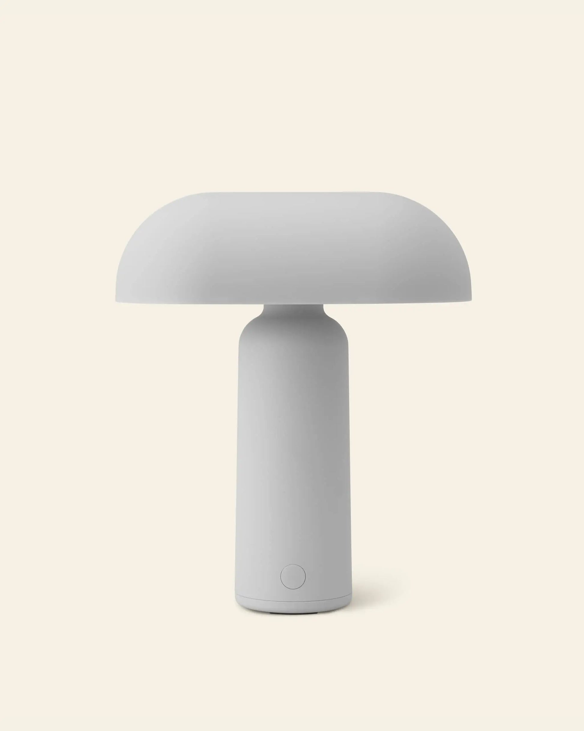 The Normann Copenhagen Porta Table Lamp features a minimalist, modern design with a mushroom-shaped shade and cylindrical base, showcased against a plain, light beige background.