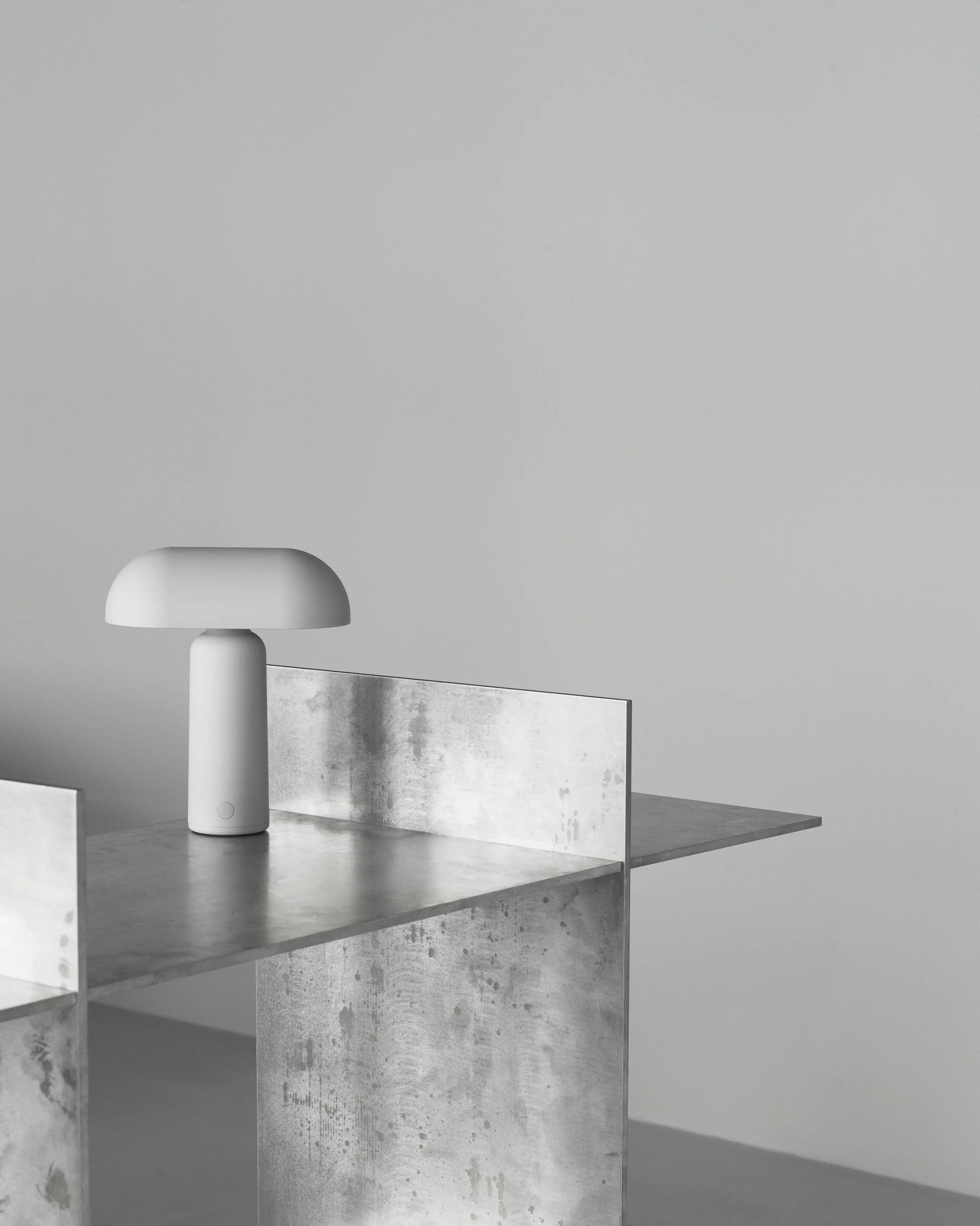 The Normann Copenhagen Porta Table Lamp, featuring a minimalist white dome-shaped shade, sits on a metallic industrial table against a plain light gray background, highlighting the modern style of Normann Copenhagen.