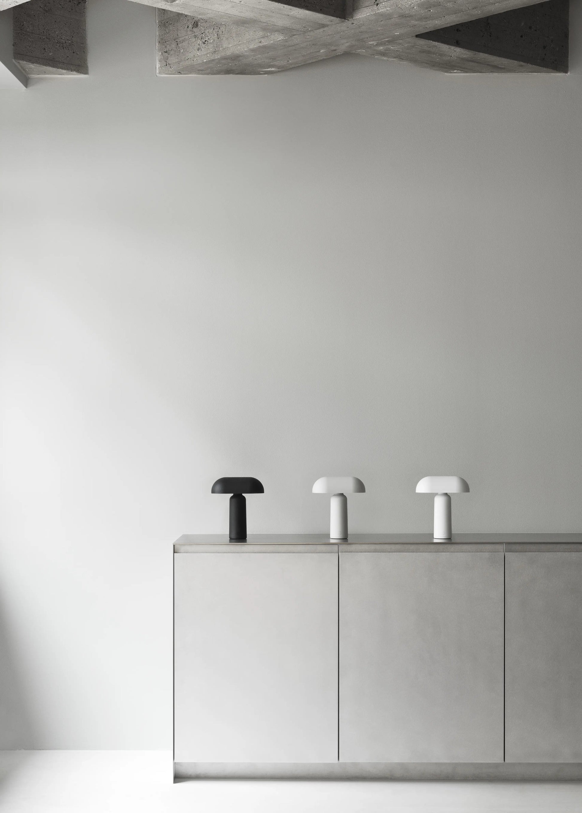 A minimalist room features a concrete ceiling, light gray walls and cabinets, and three Normann Copenhagen Porta Table Lamps in black, light gray, and white arranged on top of the cabinet.