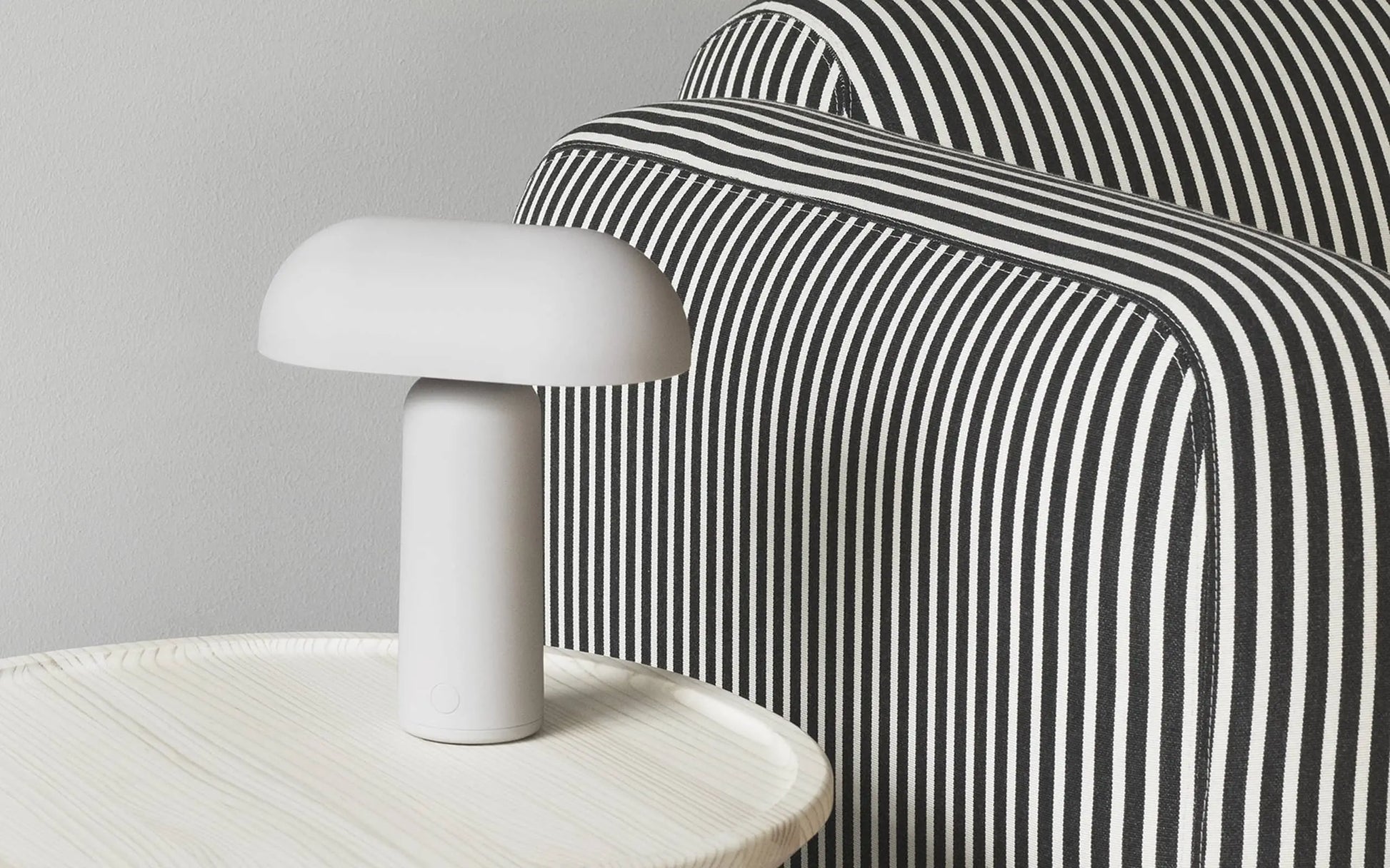 The Normann Copenhagen Porta Table Lamp in white sits on a round, light-colored table beside a black-and-white striped chair, showcasing minimalist, contemporary design by Normann Copenhagen.