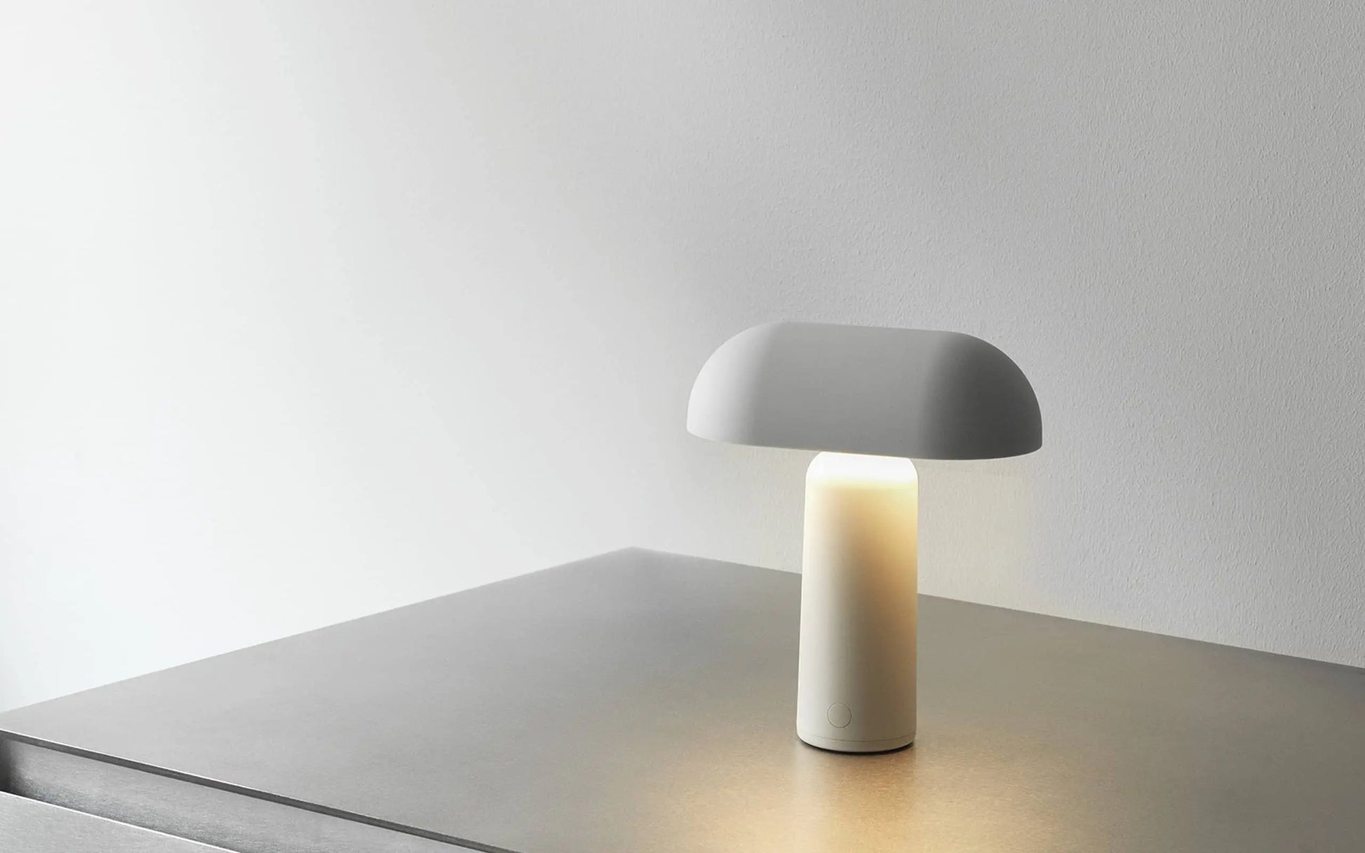 The Normann Copenhagen Porta Table Lamp, a modern minimalist piece with a rounded shade, sits on a smooth gray surface against a plain white wall, casting soft, warm light.