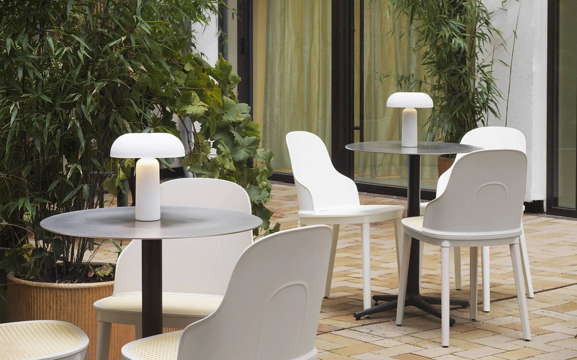 An outdoor patio with round tables and white chairs, each table featuring a Normann Copenhagen Porta Table Lamp. Green plants and large windows with yellow curtains are seen, while the floor is paved with light-colored bricks.