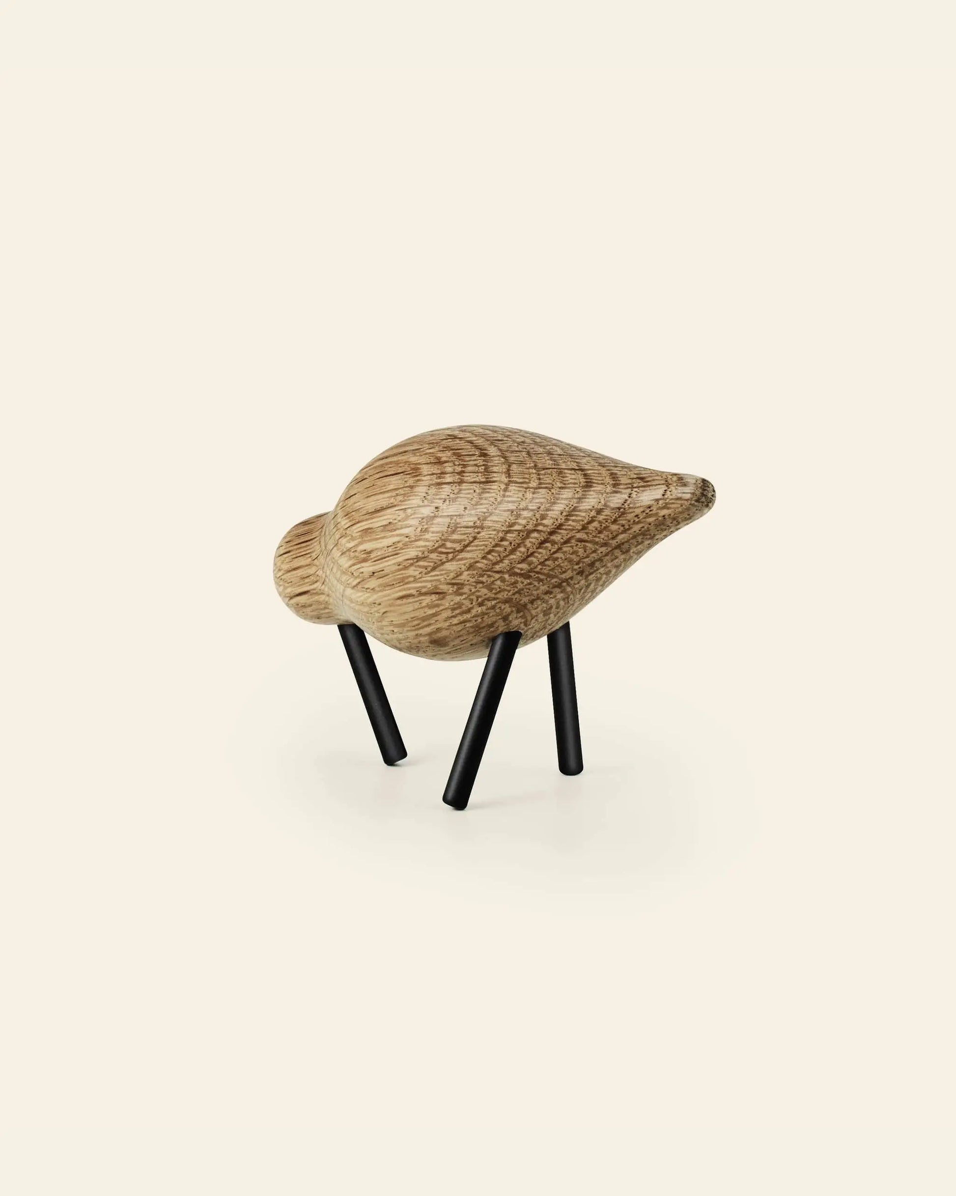 The Normann Copenhagen Shorebird Small Oak by Normann Copenhagen is a minimalist wooden bird sculpture with a rounded oak body, carved texture, and two slim black legs, displayed on a plain off-white background. Inspired by minimalist design.