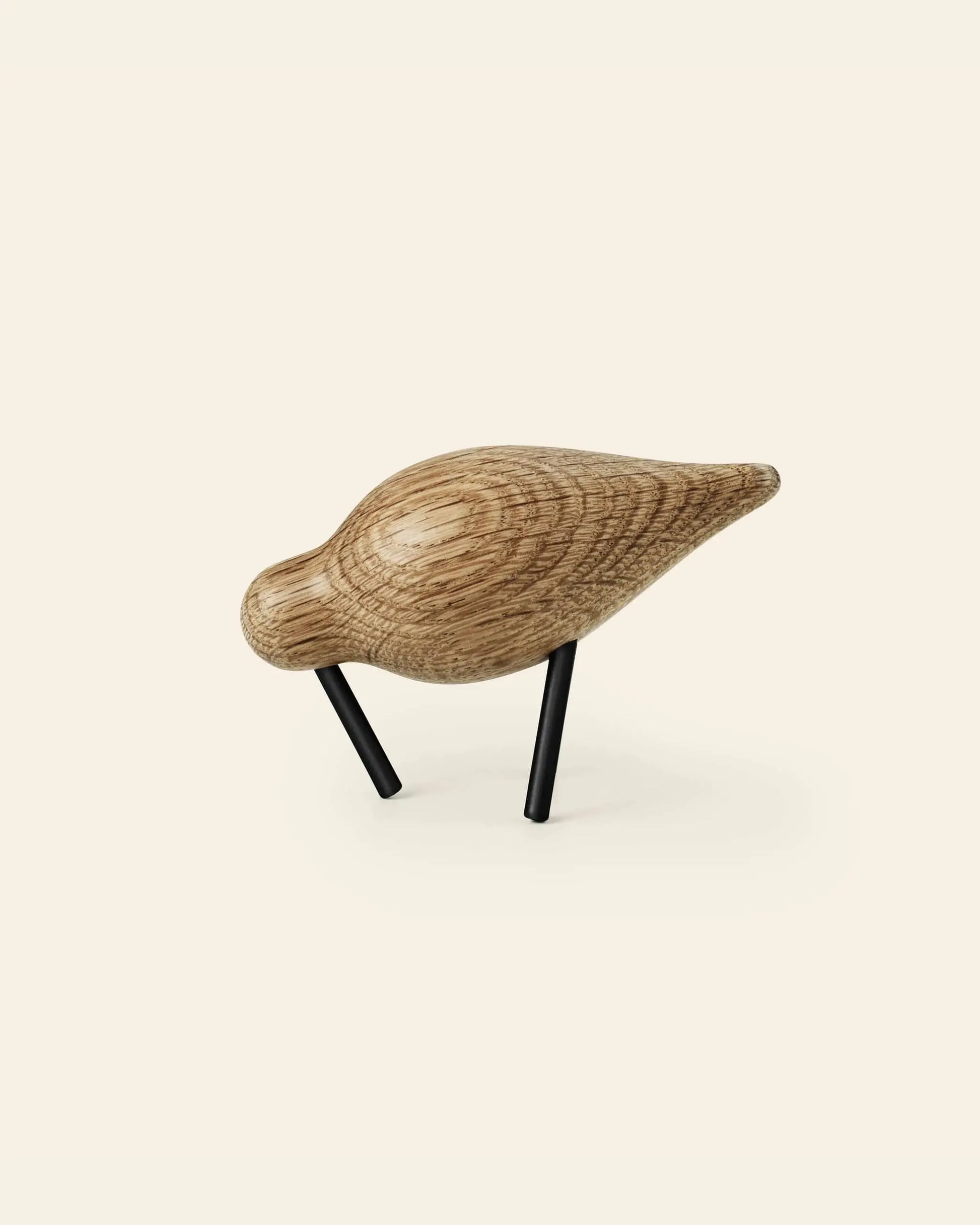 The Normann Copenhagen Shorebird Small Oak is a minimalist wooden bird sculpture crafted from oak with a smooth, natural finish and slender black metal legs, designed by Normann Copenhagen.