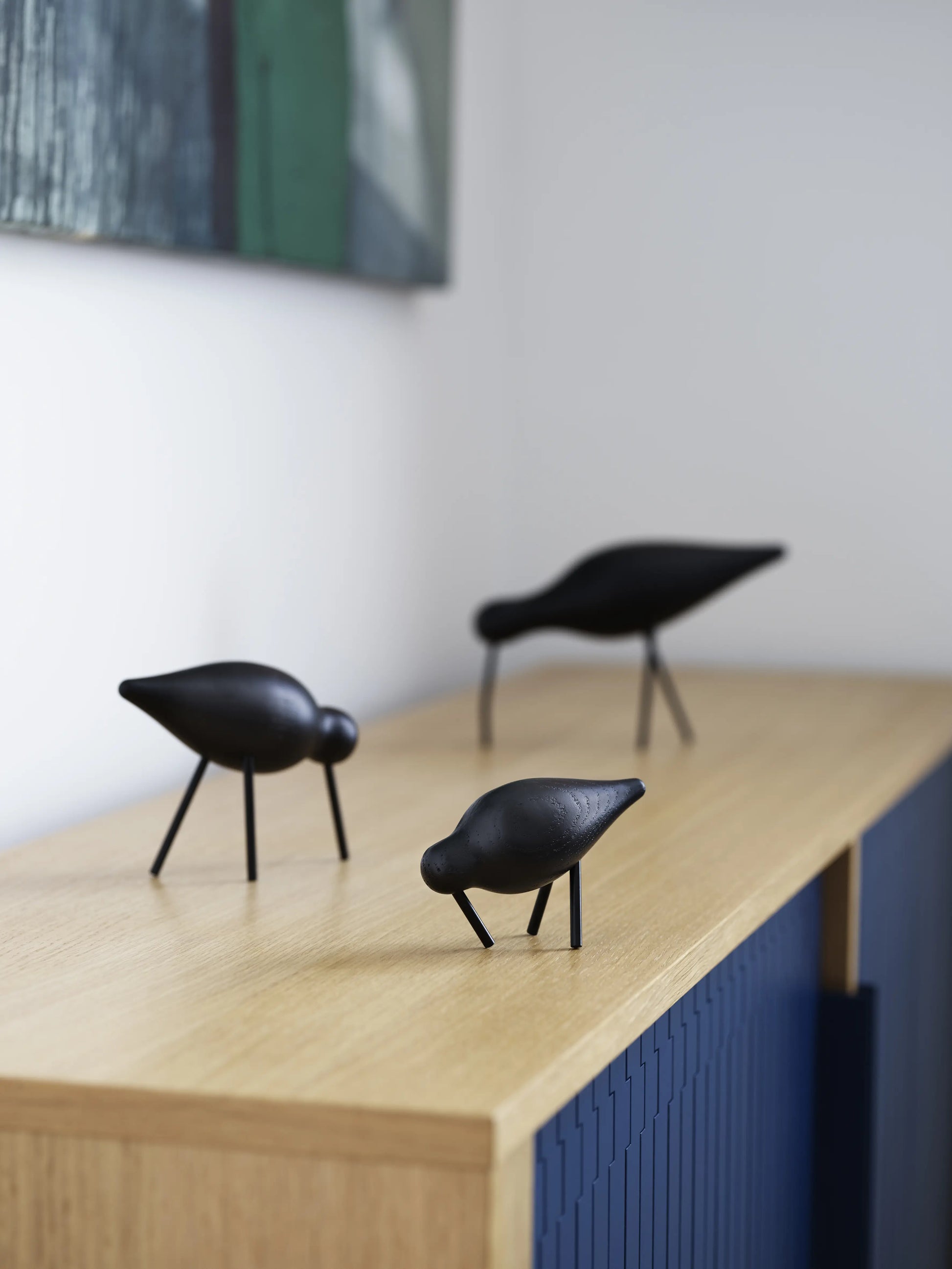 A Normann Copenhagen Shorebird Small Oak sculpture stands on a light oak surface against a white wall, with part of a framed artwork visible in the background.