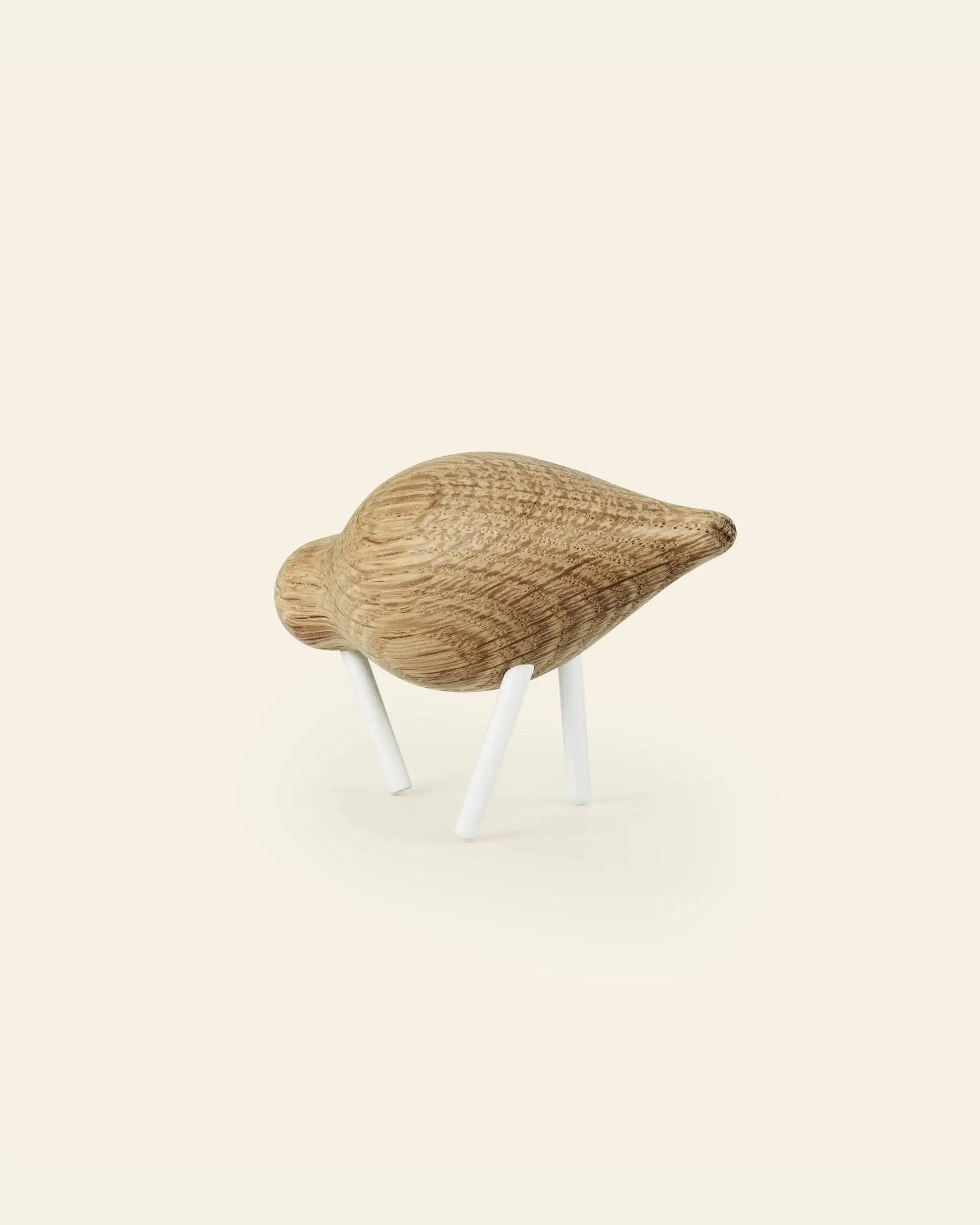 The Normann Copenhagen Shorebird Small Oak is a minimalist wooden bird sculpture with a rounded oak body, pointed beak, and two white legs, from the brand Normann Copenhagen, displayed on a light background.