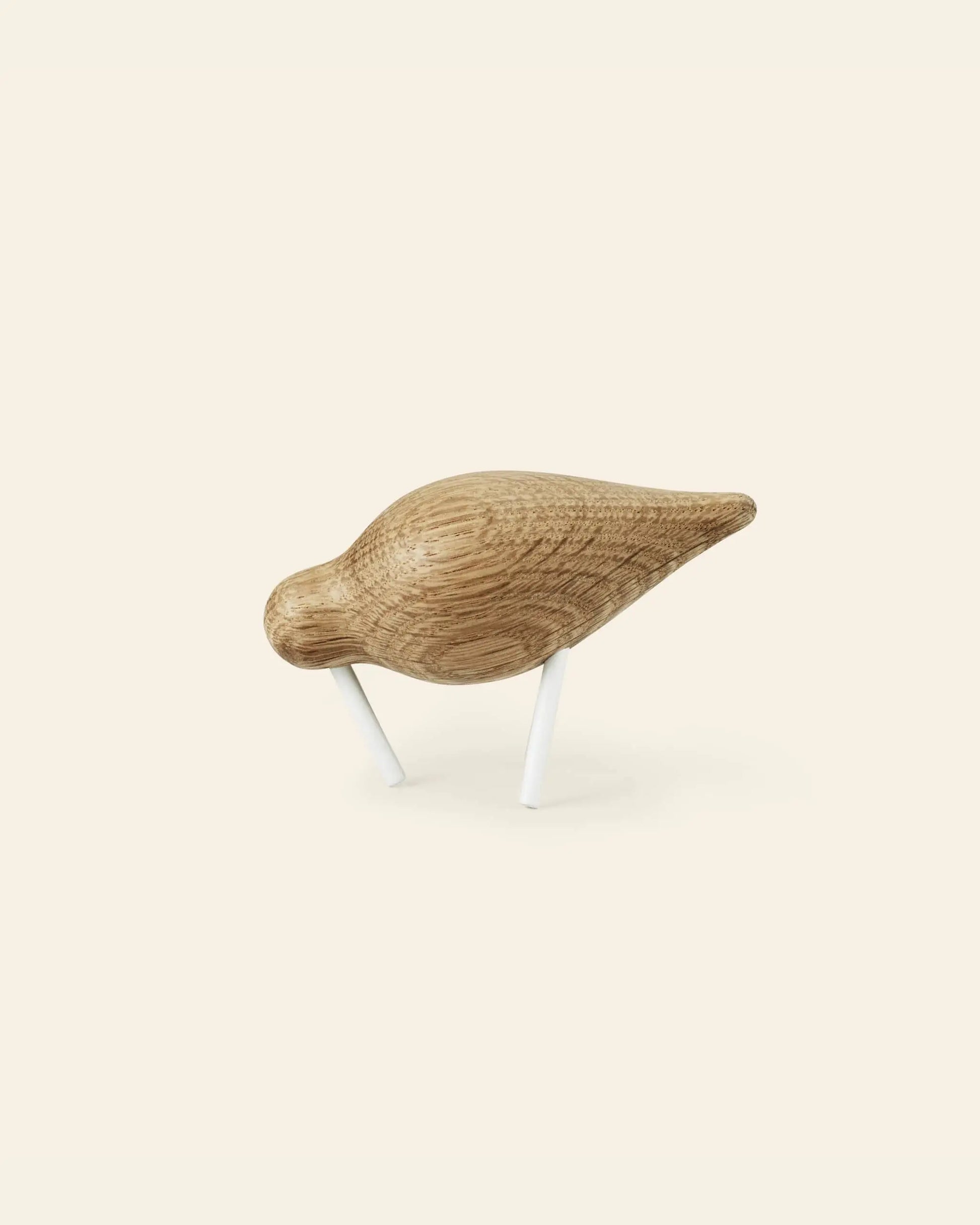 The Normann Copenhagen Shorebird Small Oak, a minimalist oak wood bird sculpture with a smooth, rounded body and two white legs, stands as if pecking against a plain light background.